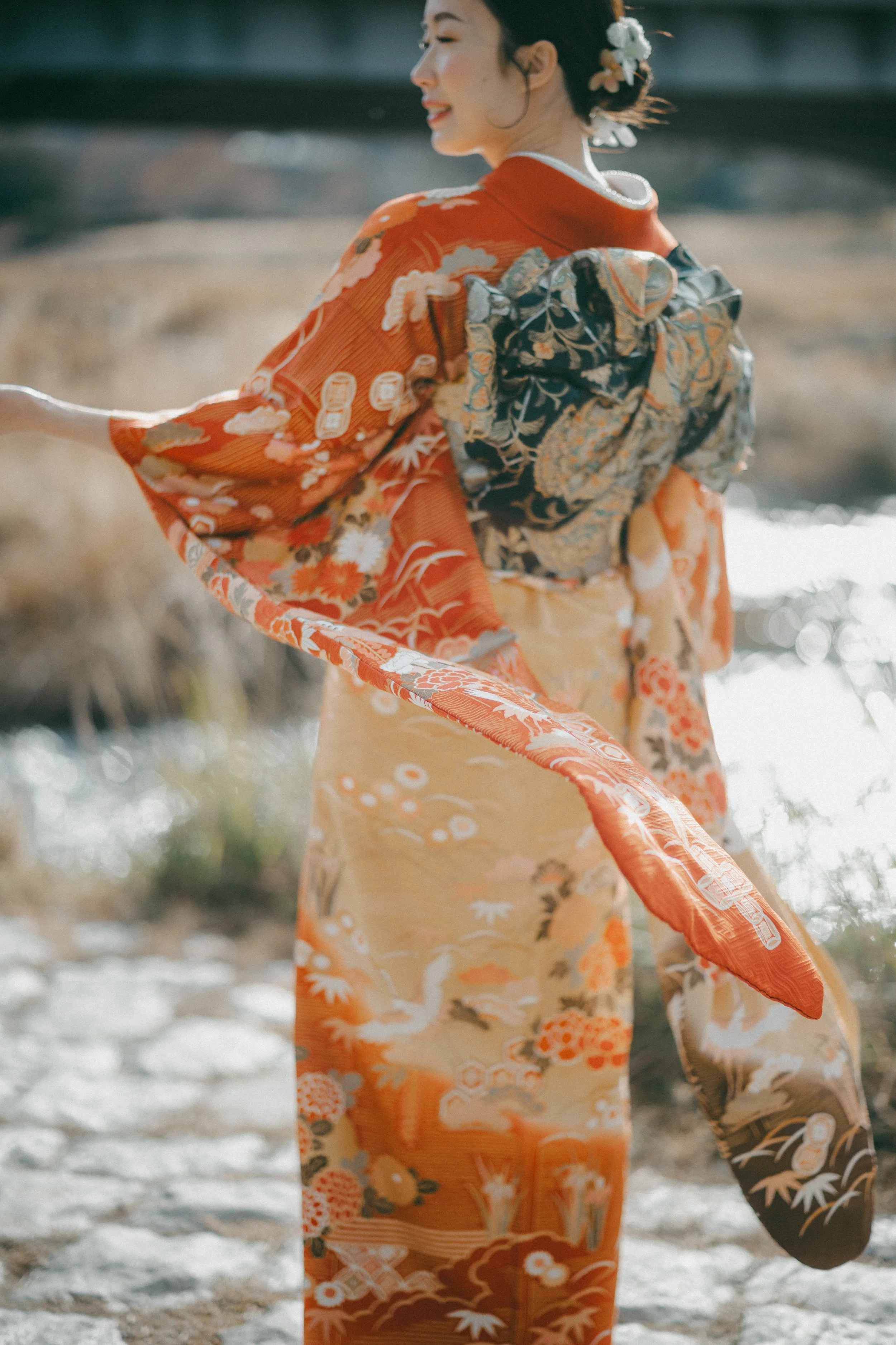 A woman wearing a traditional Japanese kimono with floral and crane patterns stands outdoors near a body of water, smiling and looking to her left.