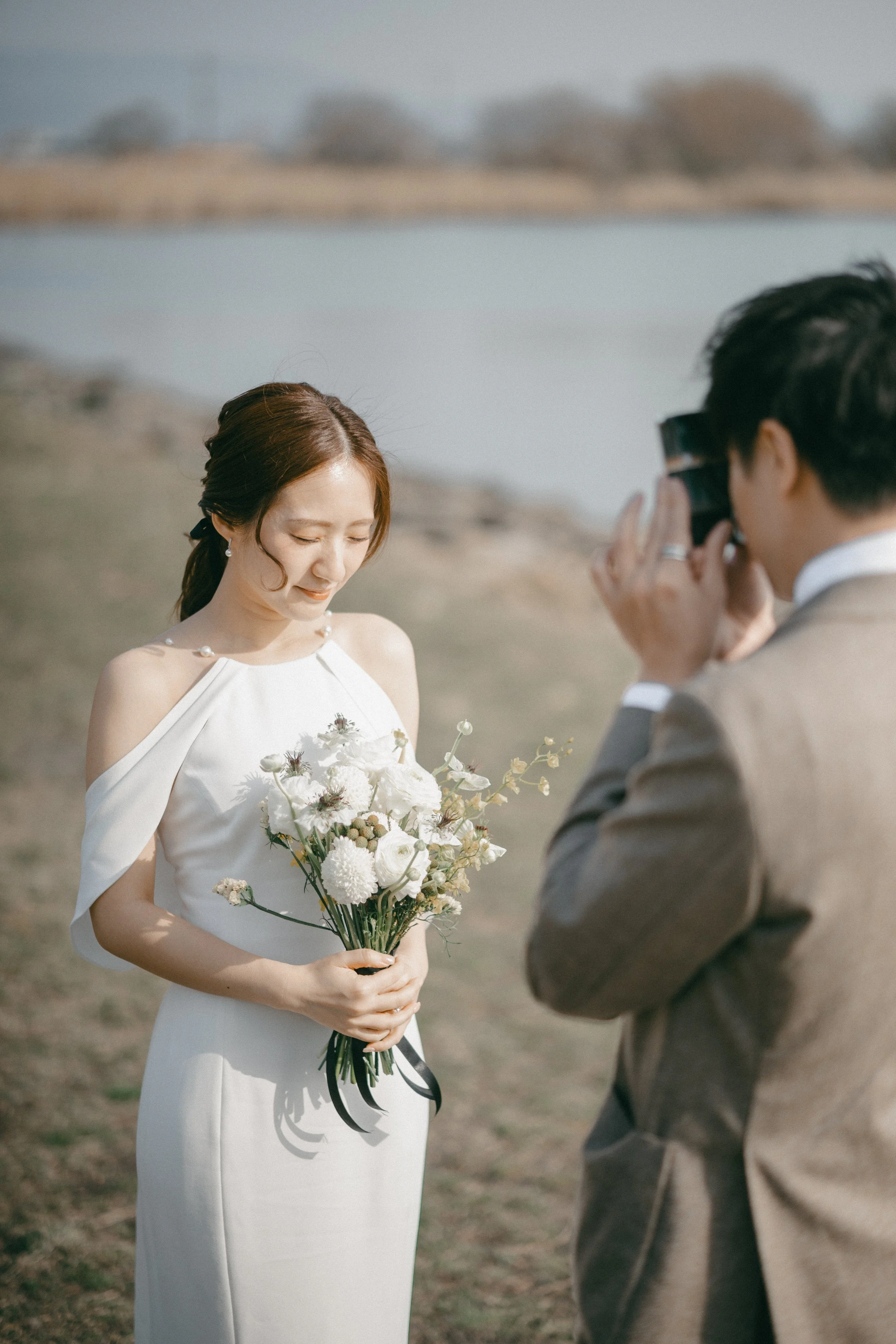 A woman in a white wedding dress holding a bouquet of white flowers, smiling softly as a man takes her photograph by a lake or river.