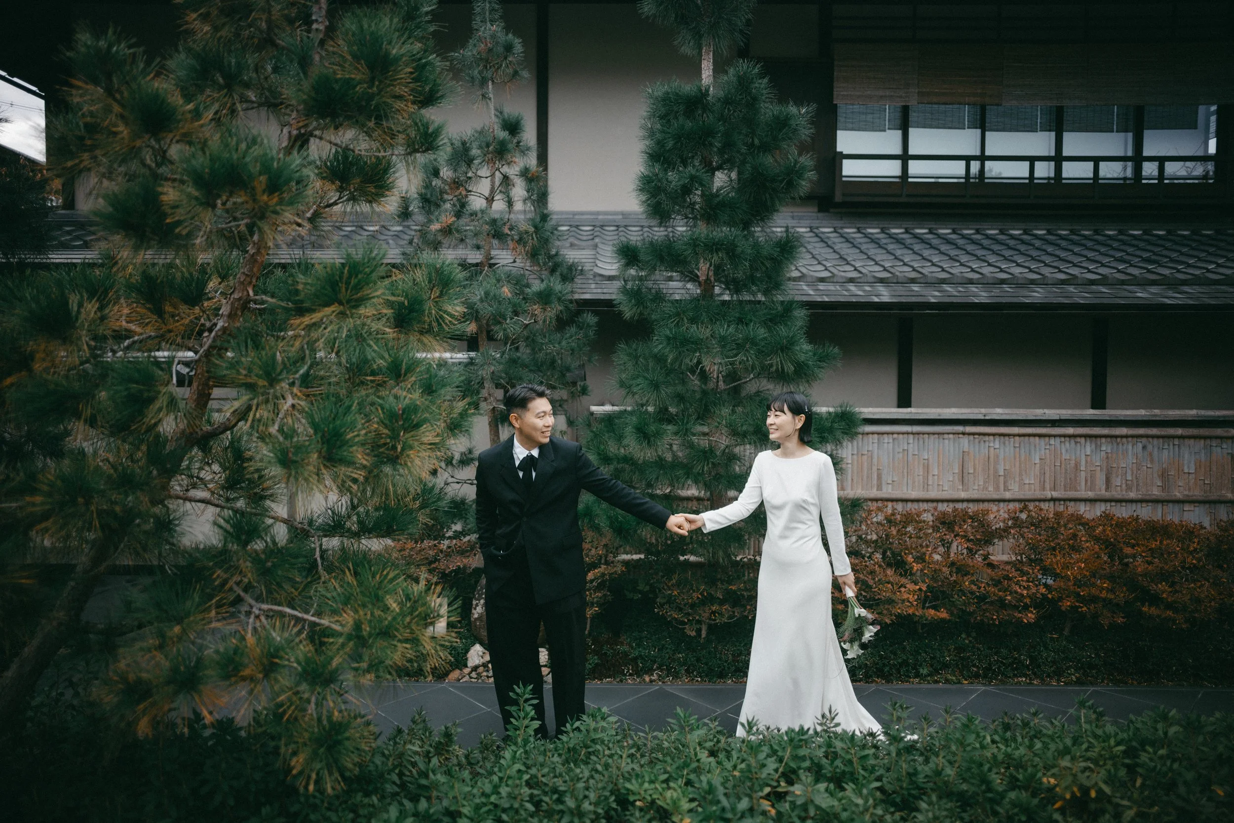 A bride and groom holding hands and smiling at each other outdoors with a large tree and traditional building in the background.