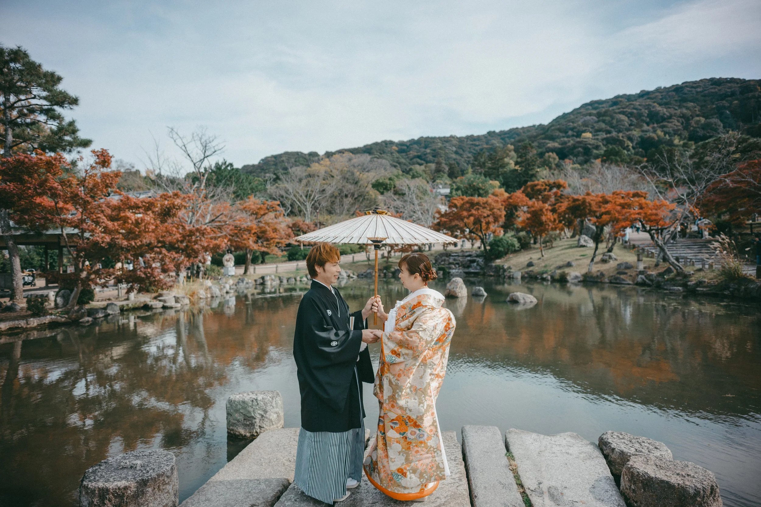 A couple dressed in traditional Japanese wedding attire exchanging vows beside a pond with autumn-colored trees in the background. The groom holds a parasol over the bride during the ceremony.