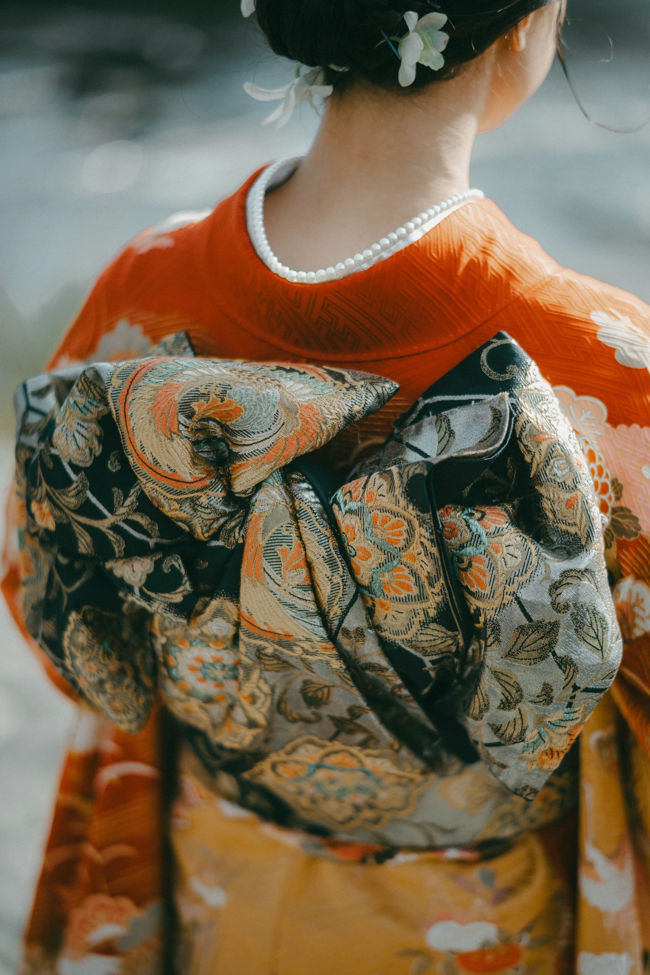 Close-up of a woman dressed in a traditional kimono with intricate brocade obi, standing outdoors. She has a string of pearls around her neck and white flowers in her updo hairstyle.