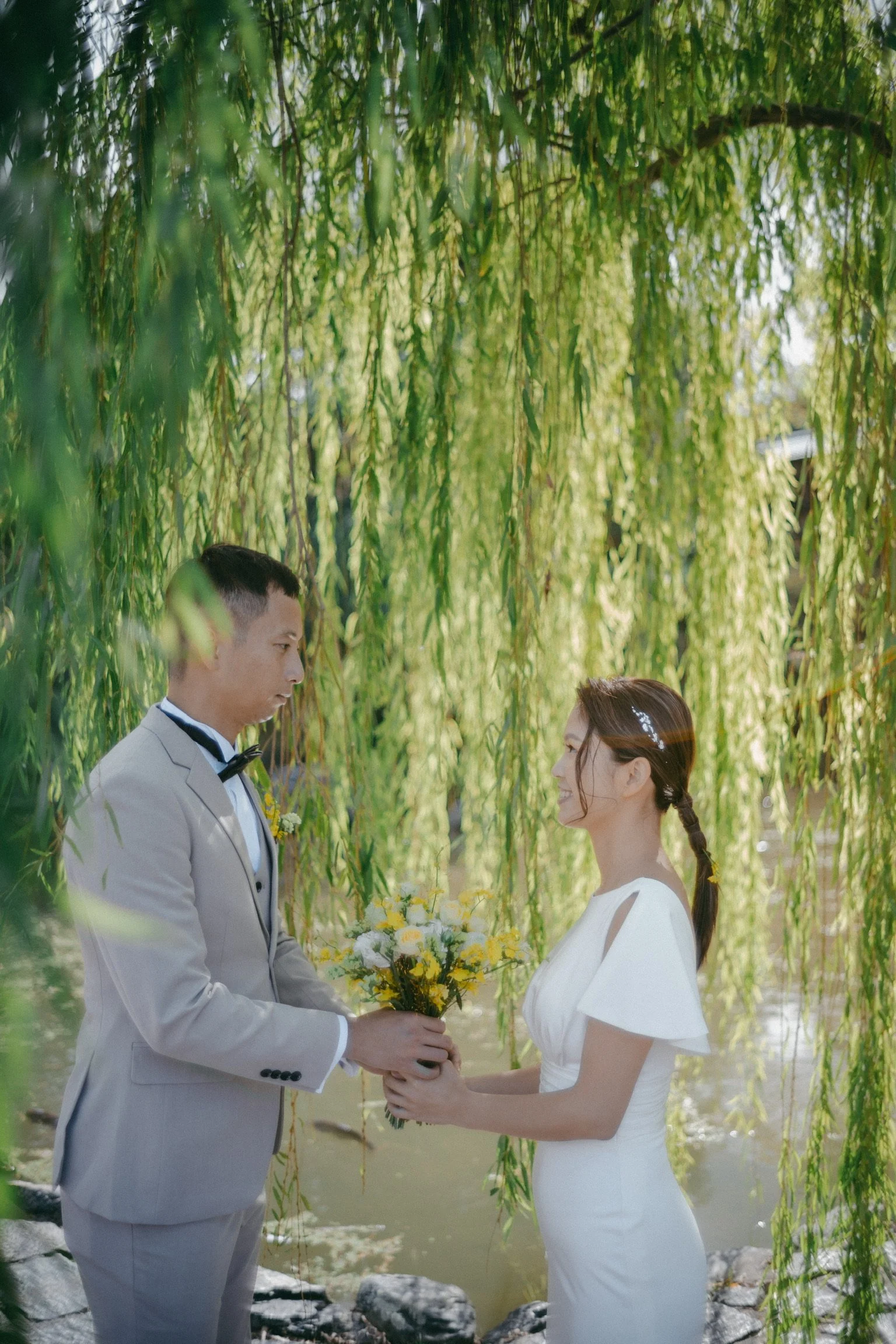 A bride and groom exchanging wedding vows under hanging green branches near a pond.