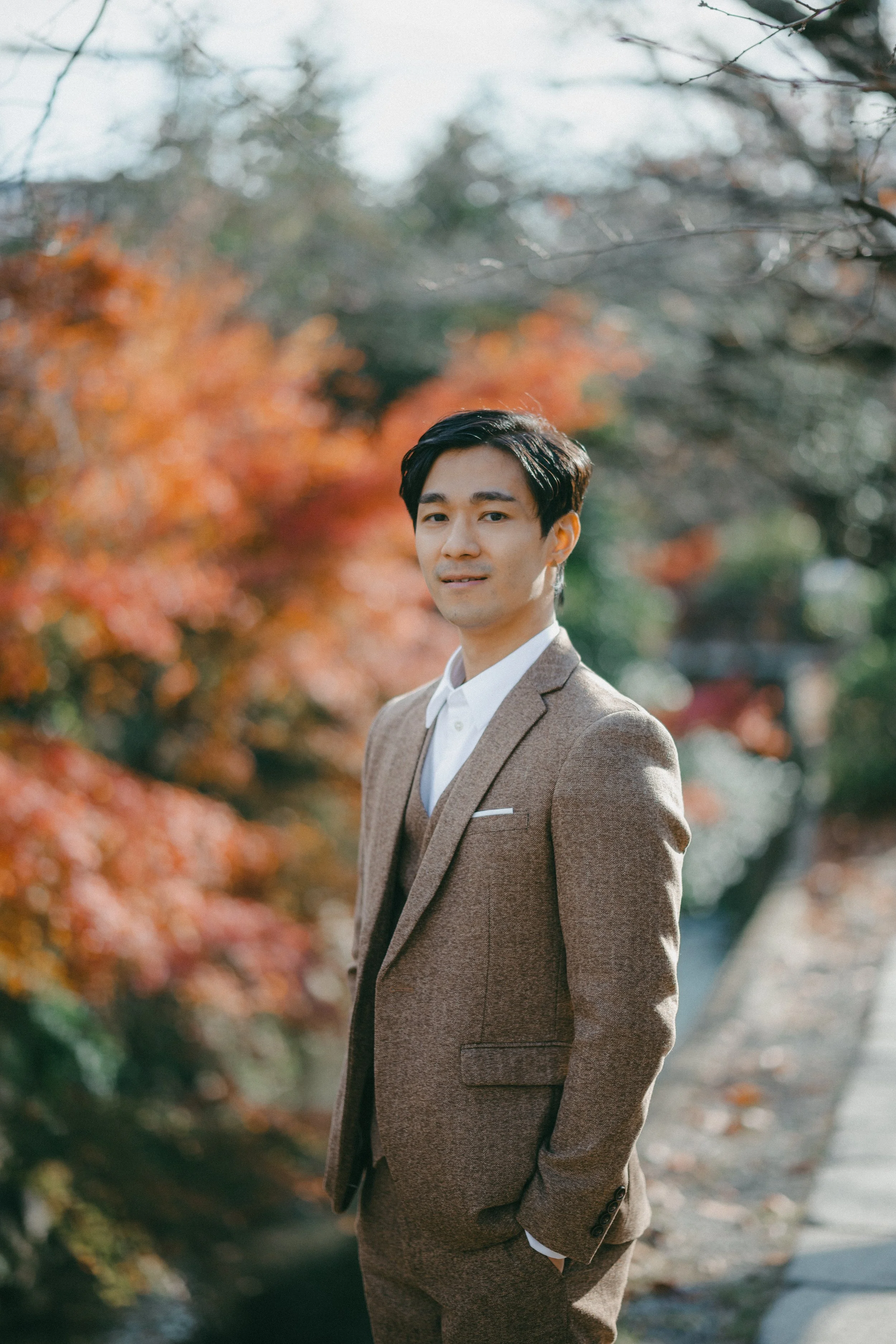 A man in a brown suit standing outdoors with autumn-colored trees in the background.