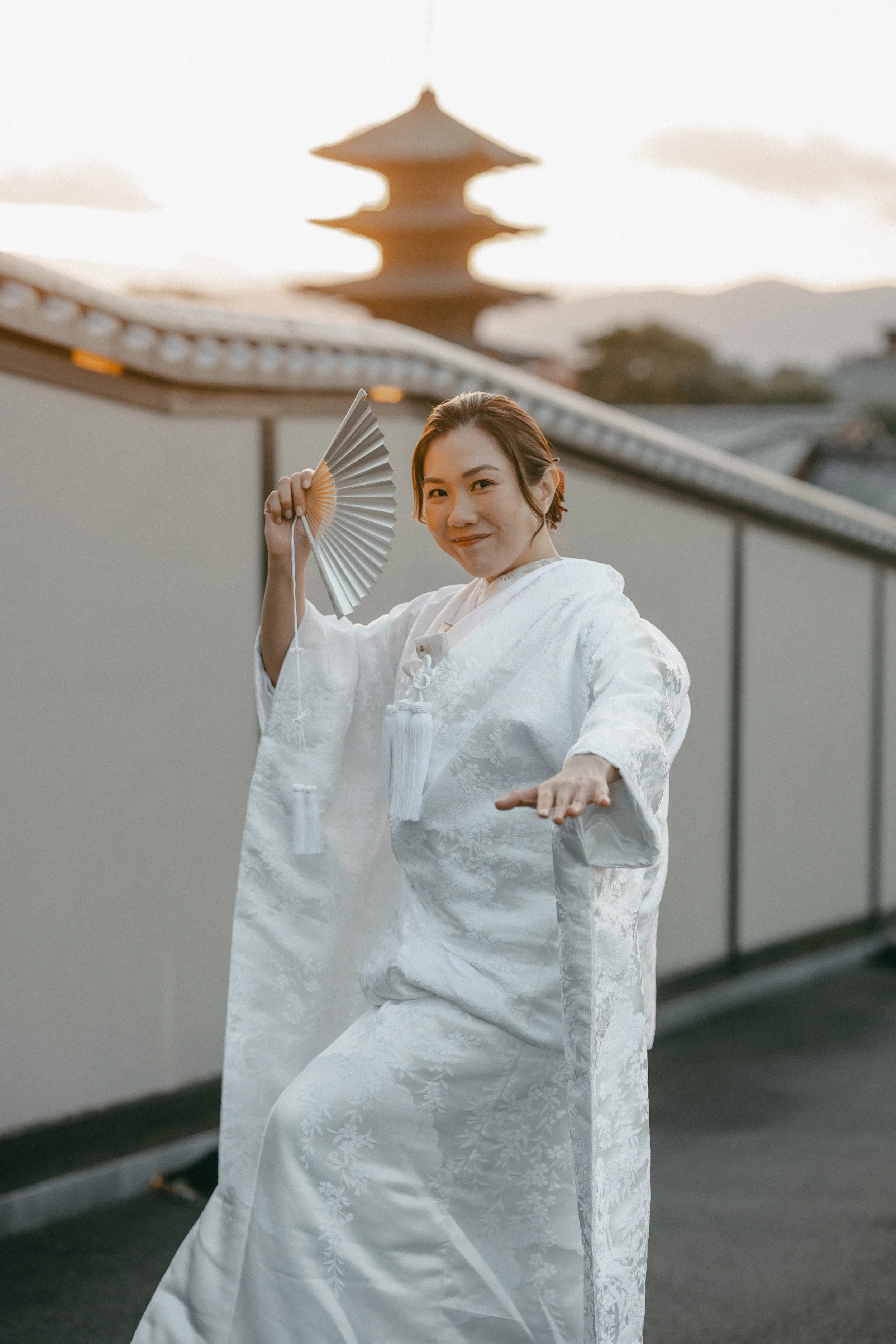 A woman dressed in a traditional white kimono holding a folding fan, performing a dance outdoors during sunset with a Japanese pagoda in the background.