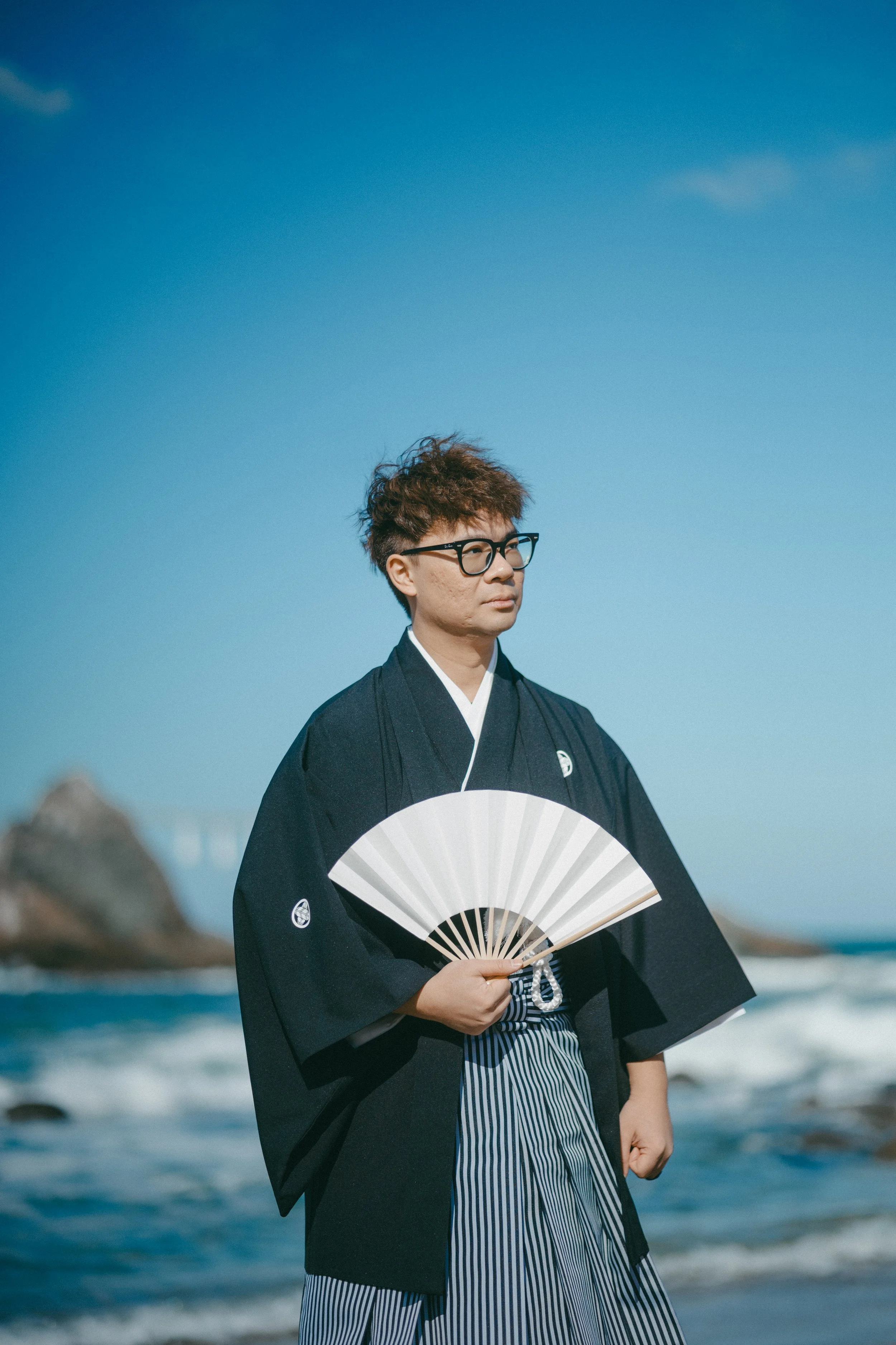 A man wearing traditional Japanese clothing, holding a paper fan, standing on a beach with rocks and water, under a blue sky.