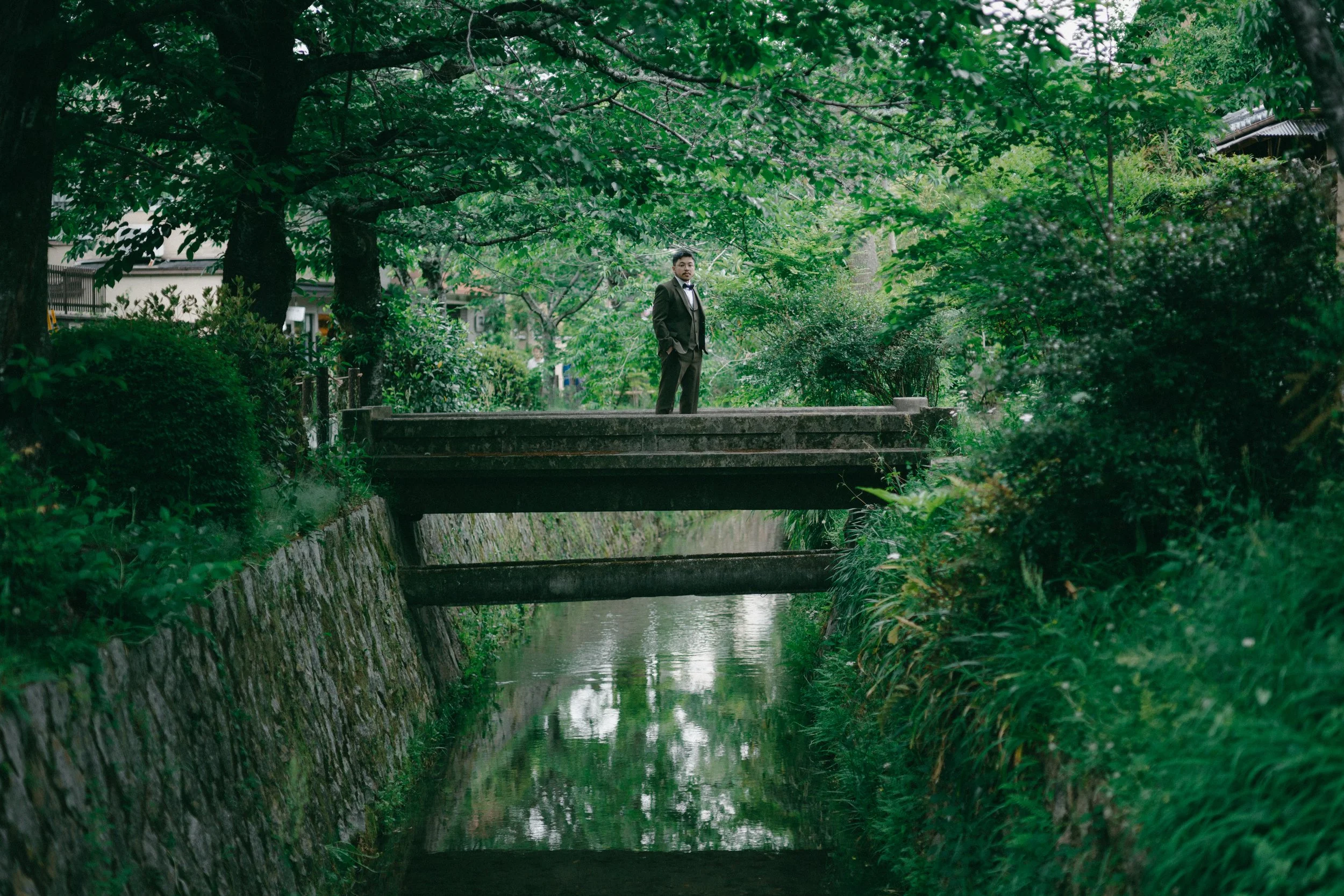 A man wearing a suit stands on a small bridge over a narrow waterway, surrounded by lush green trees and foliage.