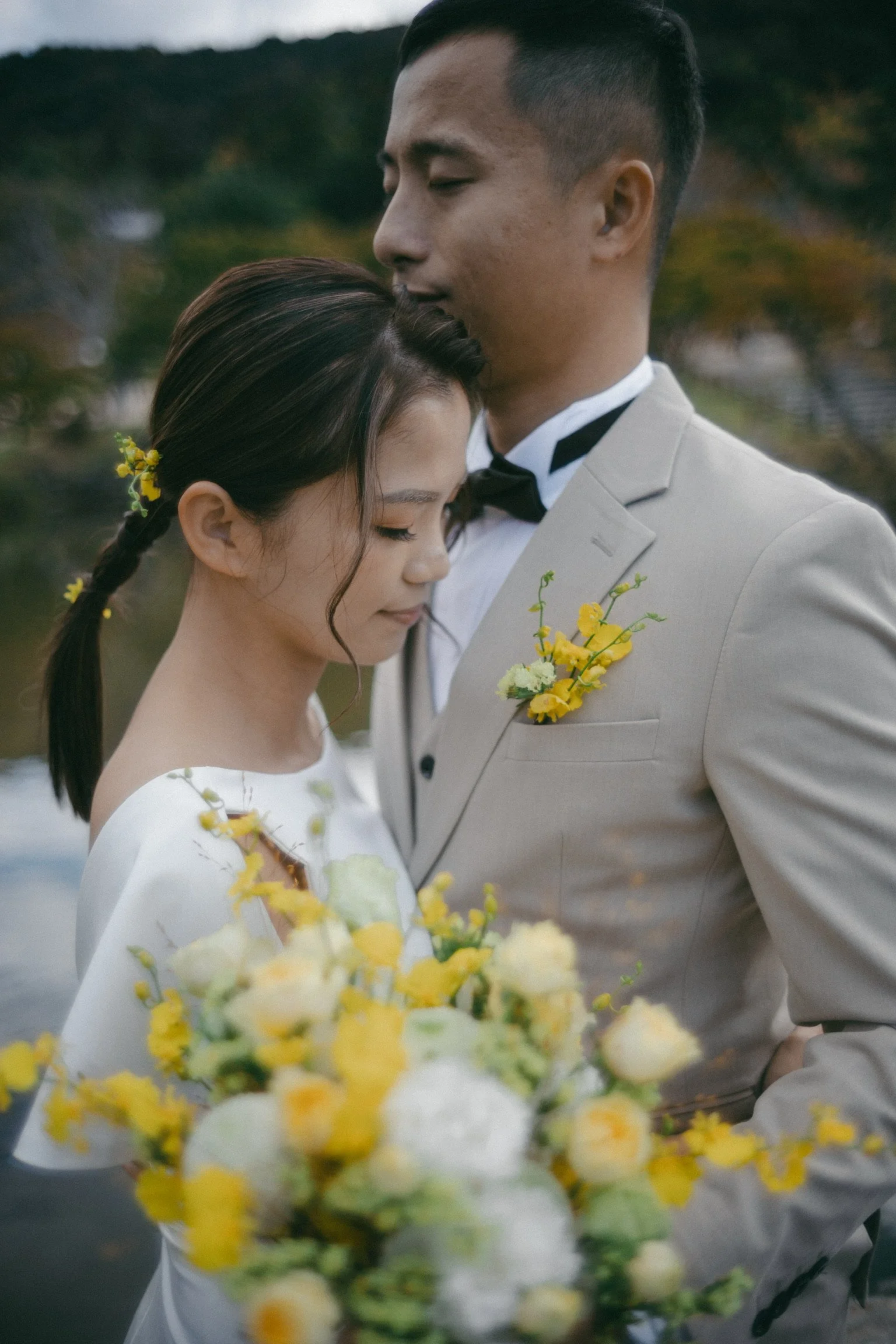A bride and groom standing close together, with heads gently touching, outdoors with a blurred natural background. The bride is holding a bouquet of yellow and white flowers, and the groom is wearing a light-colored suit with a yellow boutonniere.