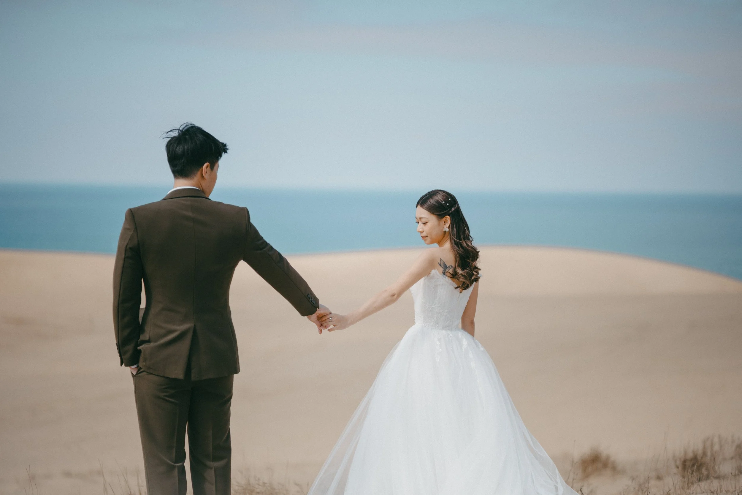 A bride and groom holding hands on a sandy beach with the ocean in the background.
