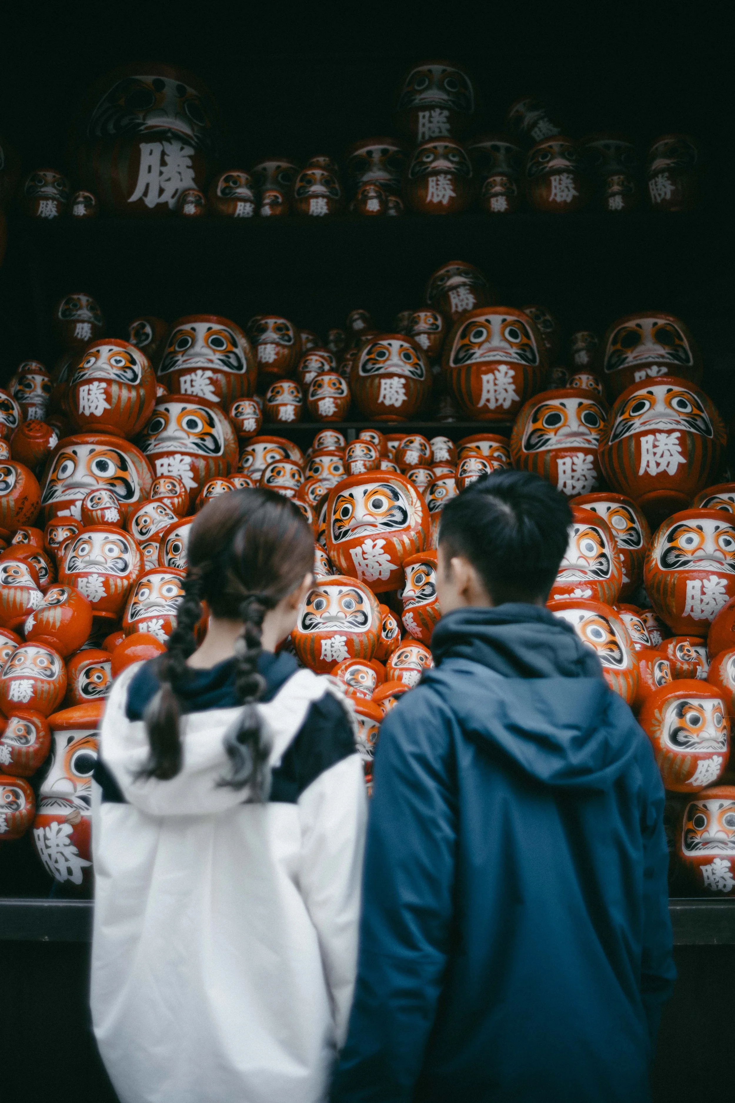 Two people with their backs turned, looking at a display of large and small Japanese Daruma dolls in red and white with black and gold details.