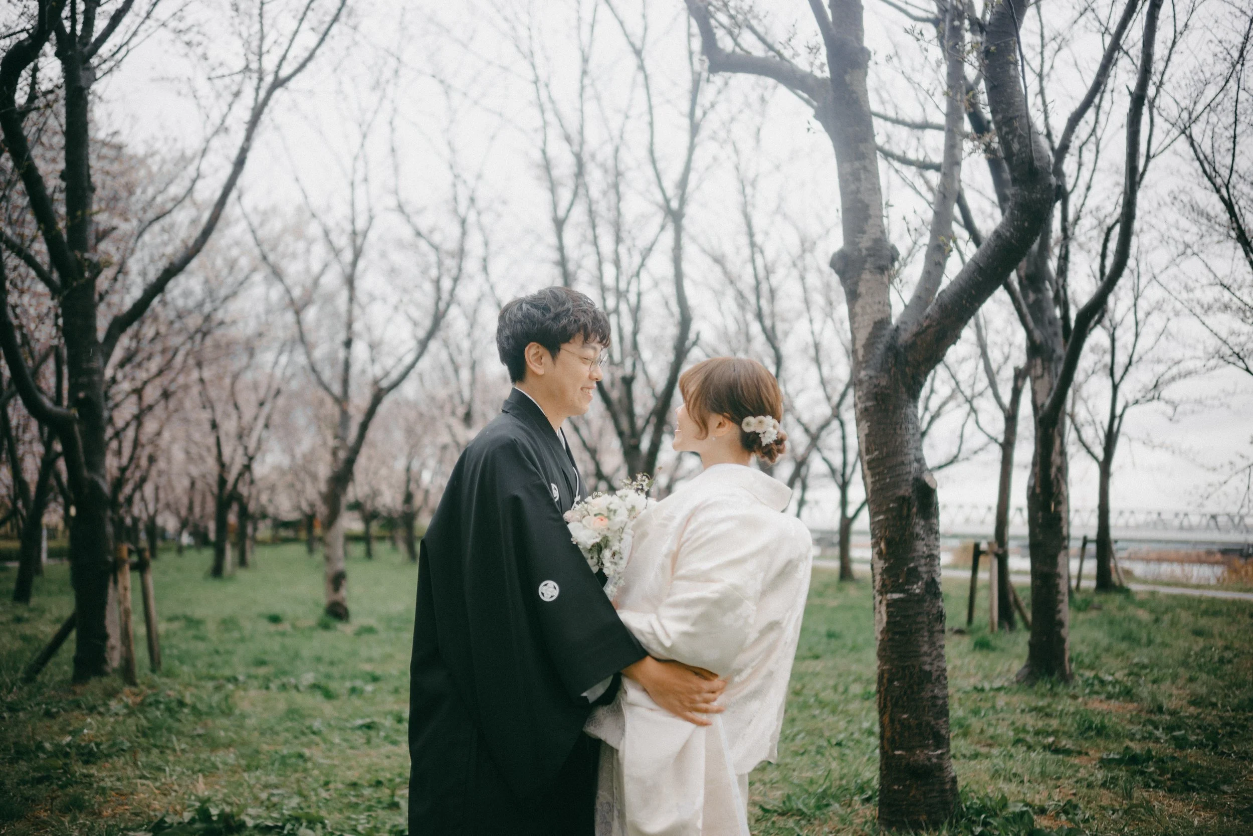 A couple in traditional Japanese wedding attire standing in a park with cherry blossom trees, holding a bouquet of flowers, and gazing at each other.