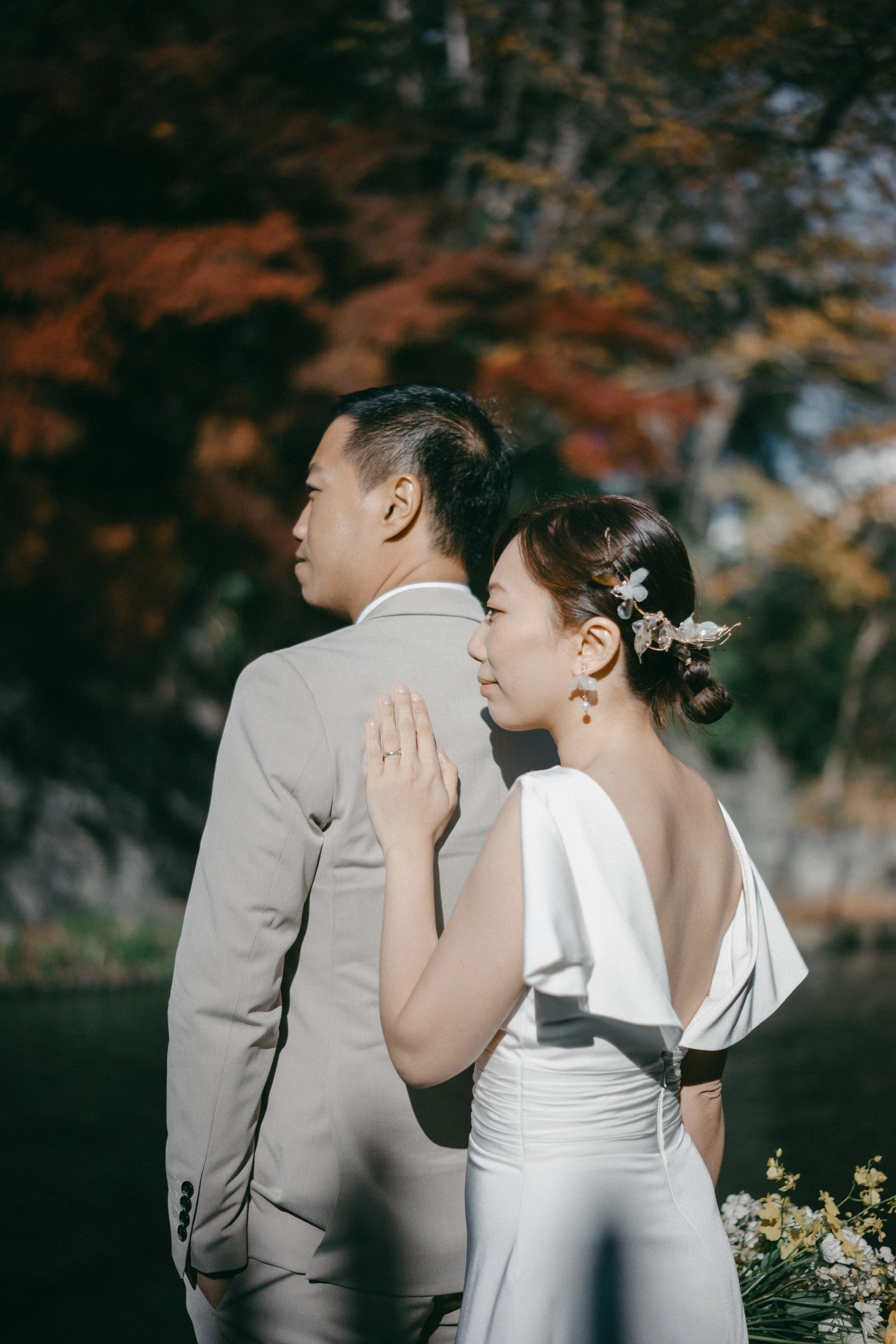 A bride and groom standing outdoors during their wedding, facing away from each other, with the bride gently touching the groom's back. The bride is wearing a white gown with off-shoulder sleeves and floral hair accessories, while the groom is in a l