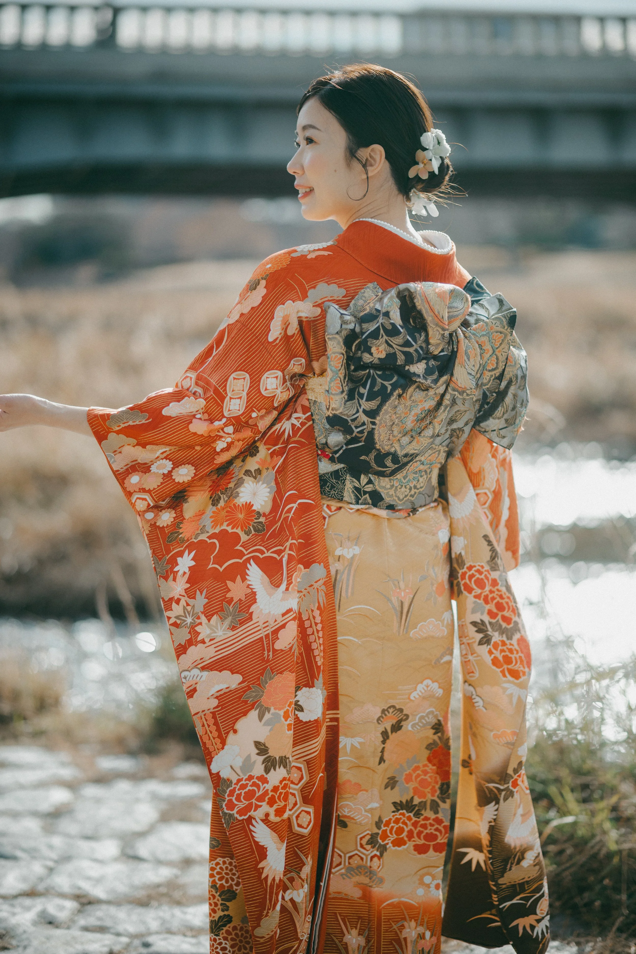 A woman wearing a traditional Japanese kimono with intricate patterns, standing outdoors near a river, with her hair adorned with flowers and a large bow on her back, smiling and looking to her left.