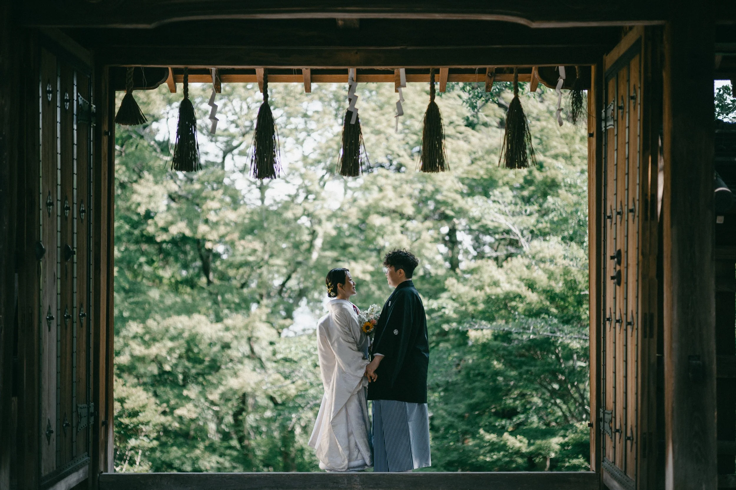 A couple in traditional Japanese wedding attire holding hands and gazing at each other, standing in a wooden temple doorway decorated with hanging tassels and wooden ornaments, surrounded by lush green trees.