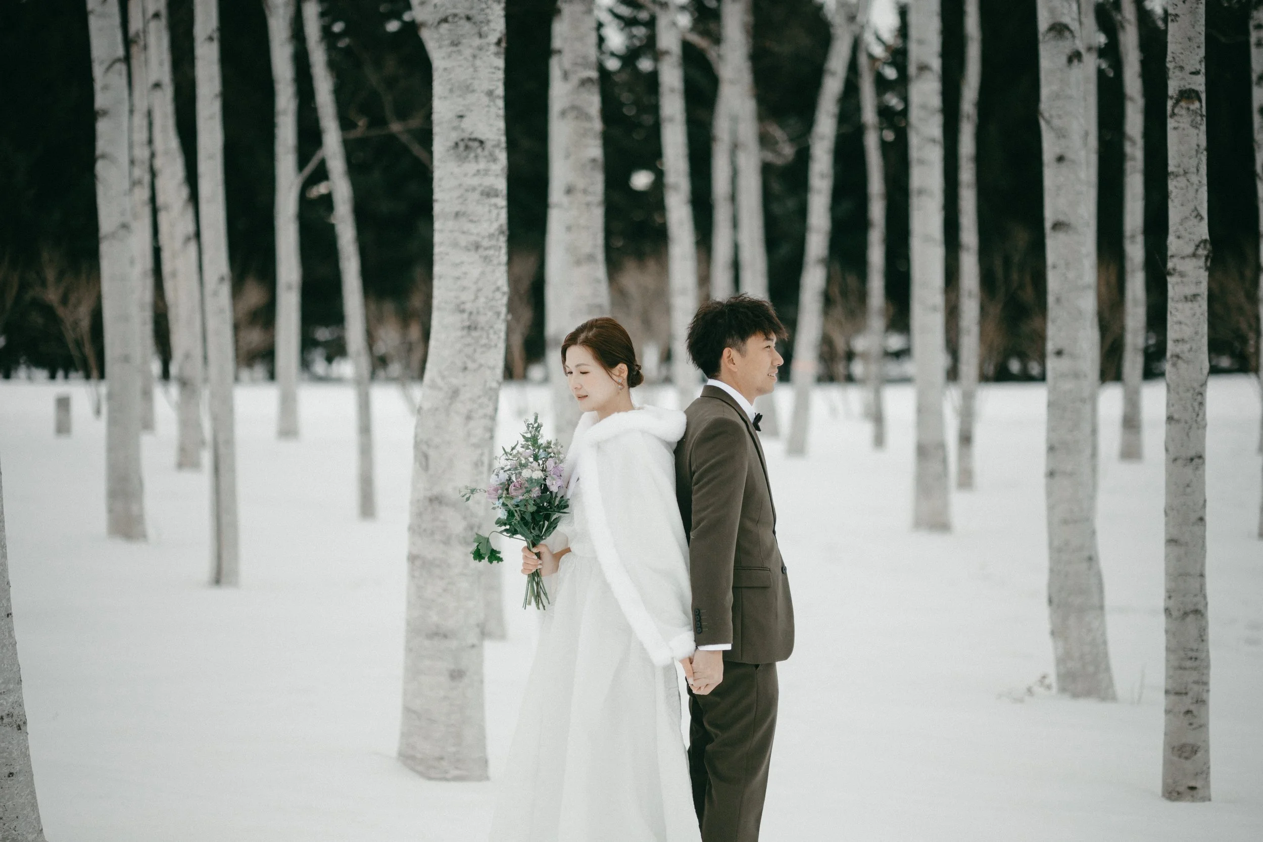 A bride and groom standing back-to-back in a snow-covered forest, holding hands, with the bride holding a bouquet of flowers.