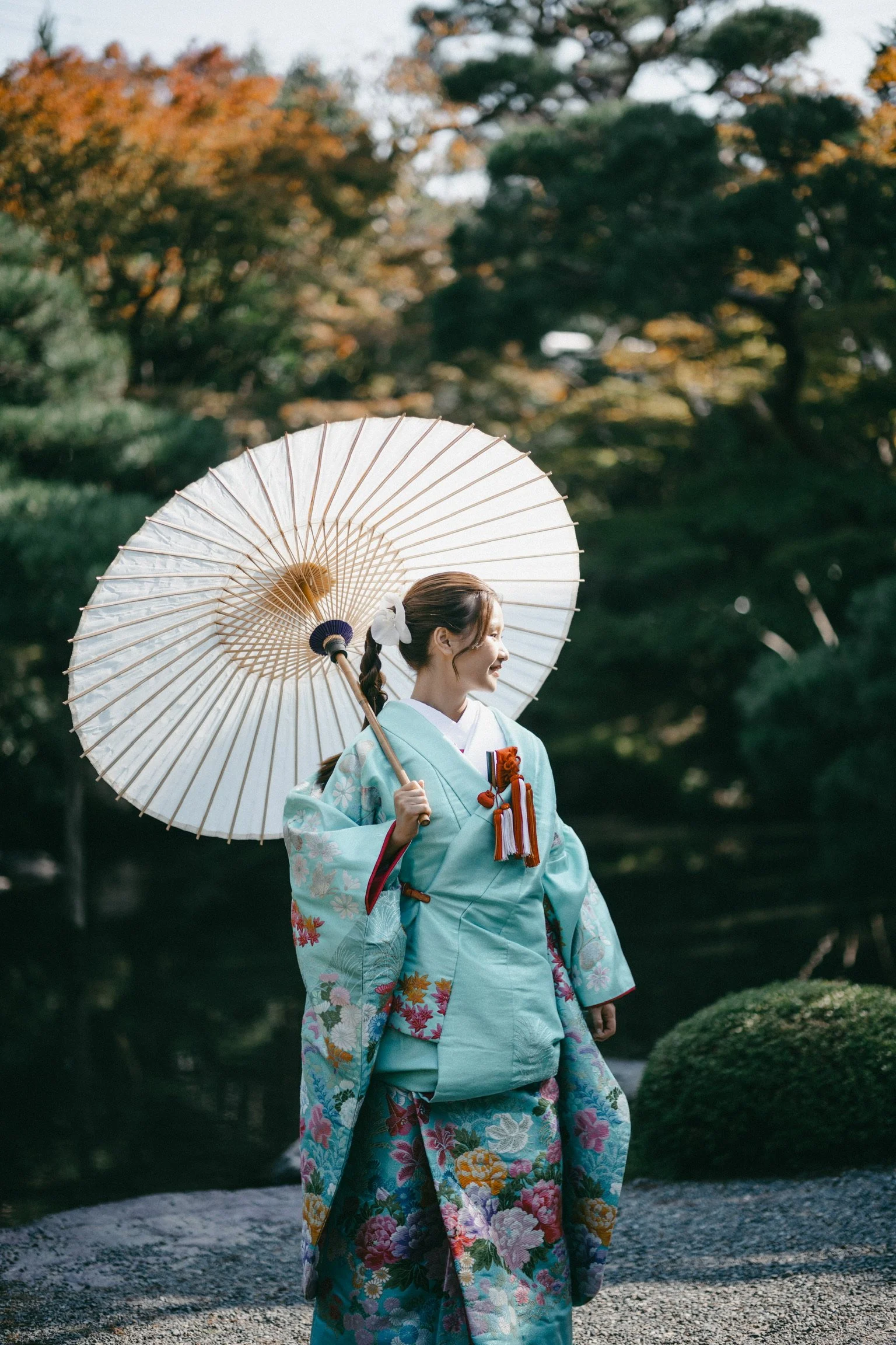 A woman dressed in a traditional Japanese kimono holding a white parasol, standing outdoors with trees and greenery in the background.
