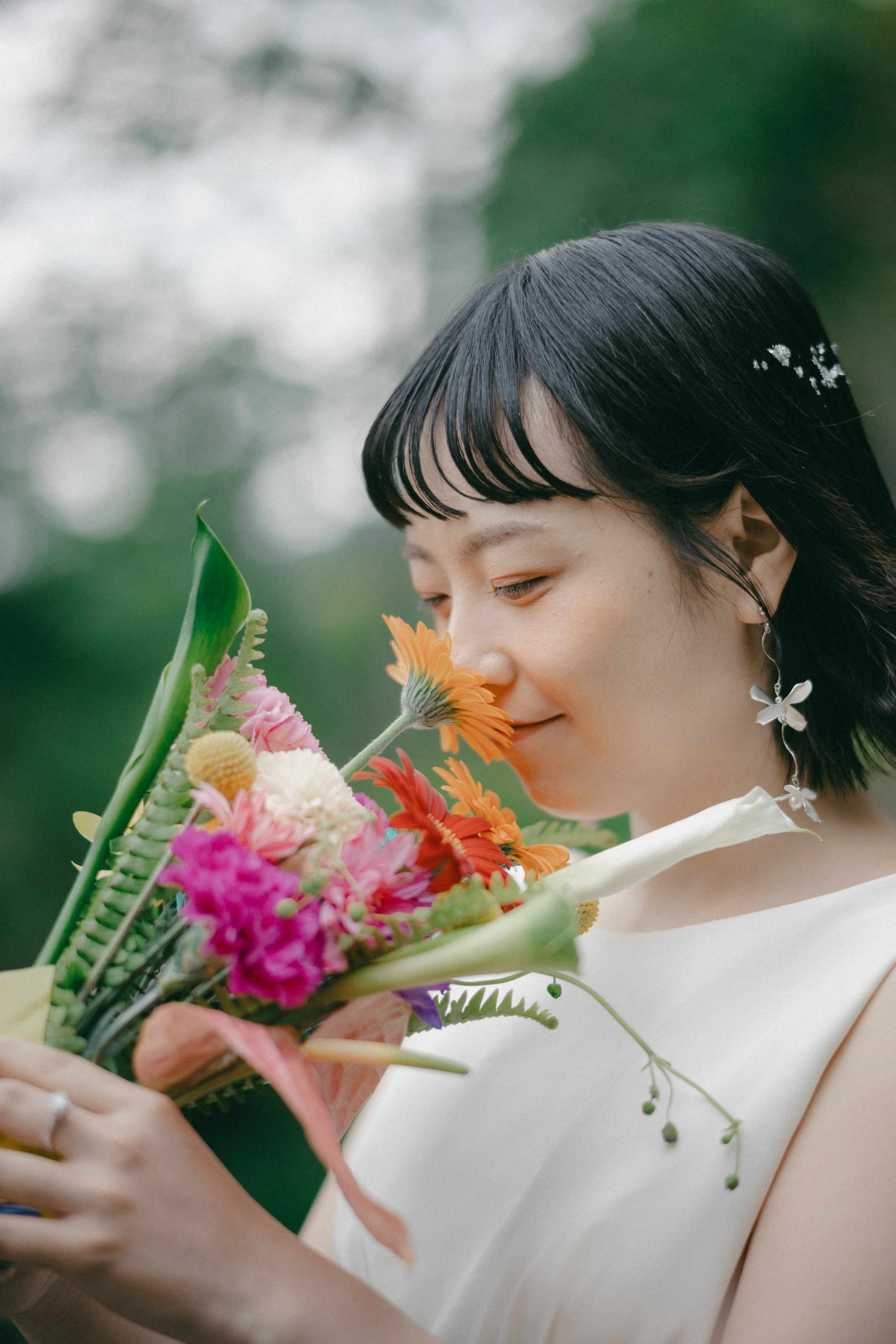 A woman with short black hair, wearing butterfly earrings and a white top, holds a colorful bouquet of flowers and is smelling them outdoors.