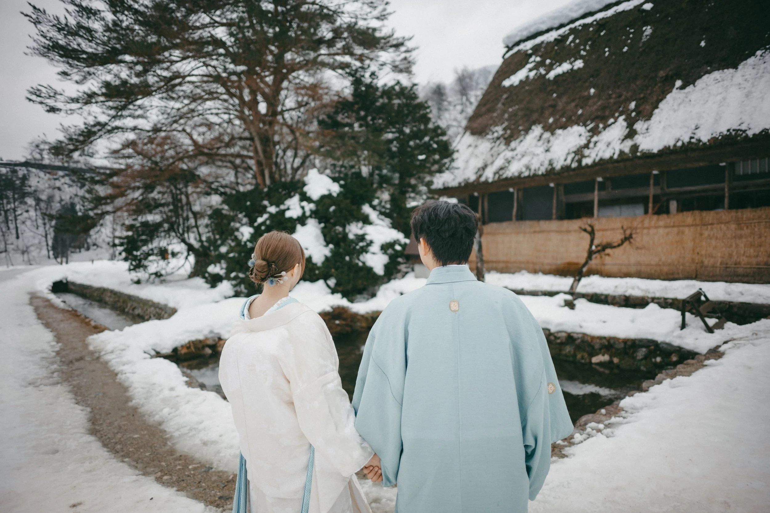 A couple in traditional Japanese wedding attire holding hands outdoors in a snowy landscape, with trees, a small stream, and a thatched-roof building in the background.