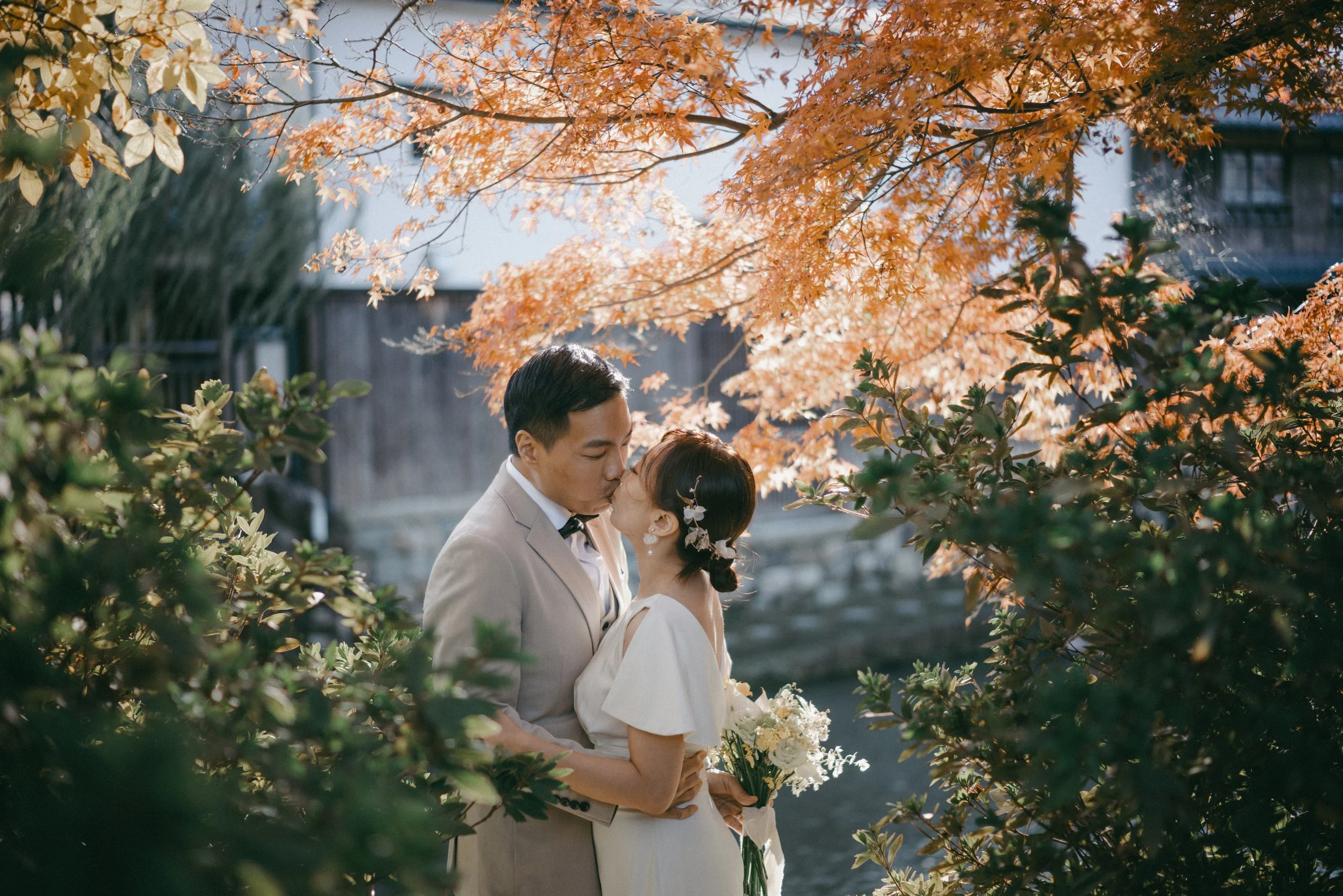 A couple dressed in wedding attire sharing a kiss outdoors surrounded by trees with autumn leaves.