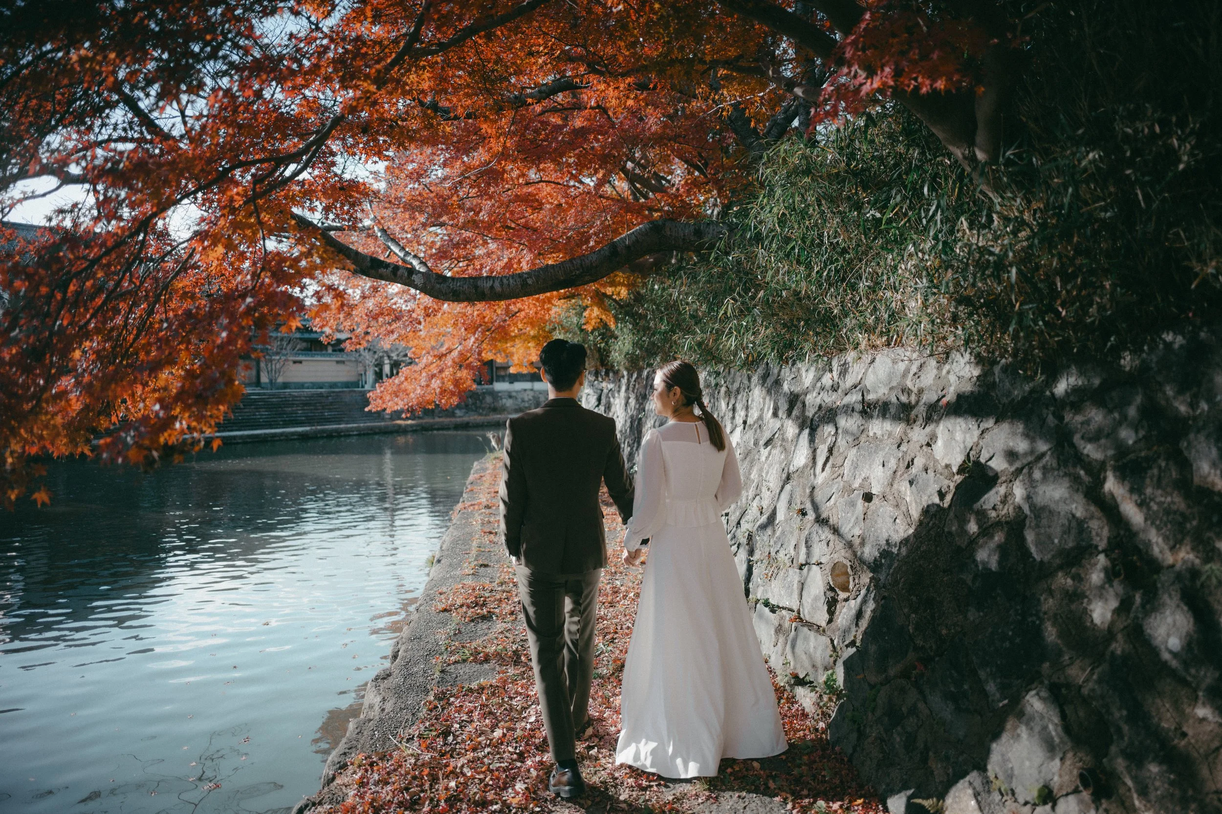 A couple dressed in wedding attire walking hand in hand along a stone path beside a calm river, with vibrant orange and red autumn leaves overhead.