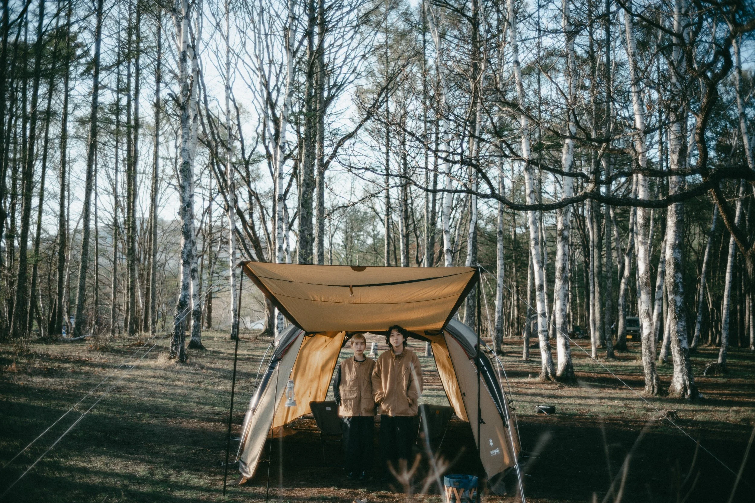 Two people standing inside a camping tent in a wooded area with tall, leafless trees and clear sky.