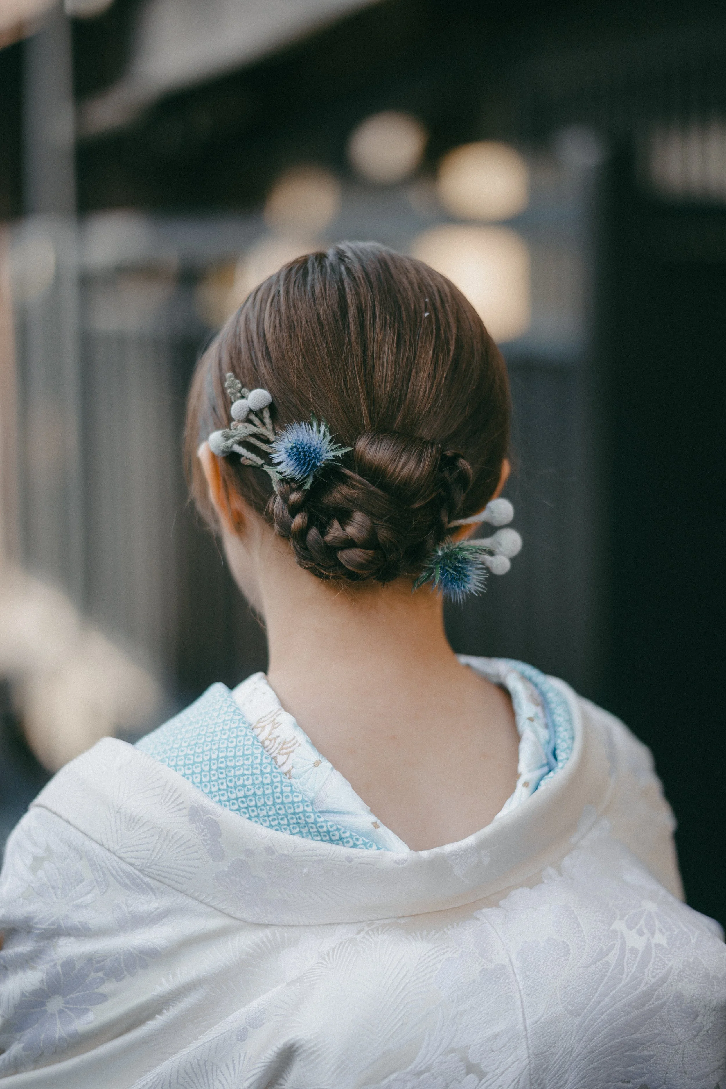 Back view of a woman with an intricate braided hairstyle adorned with blue and white floral accessories, dressed in a white kimono with light blue accents.
