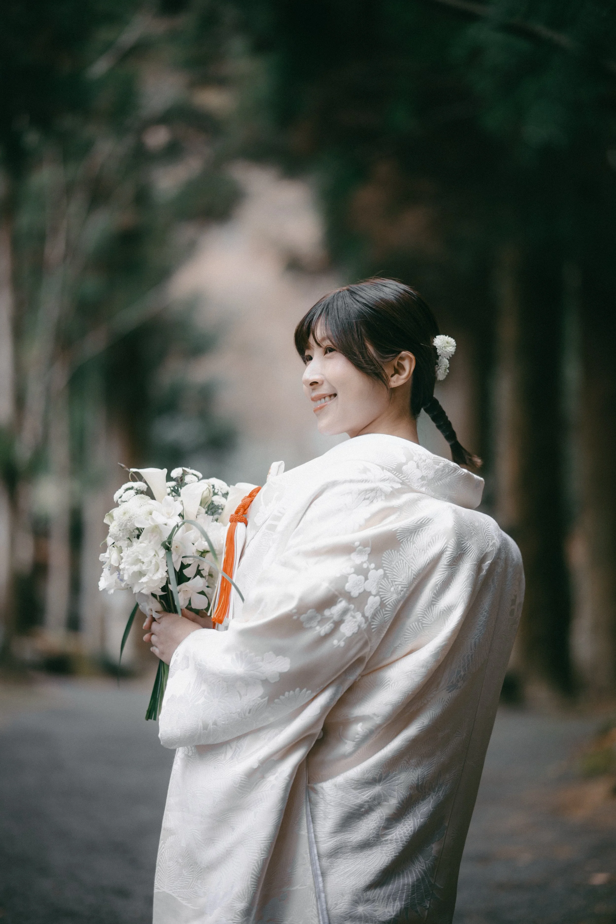 A woman in a traditional white kimono, holding a bouquet of white flowers, standing outdoors in a wooded area.