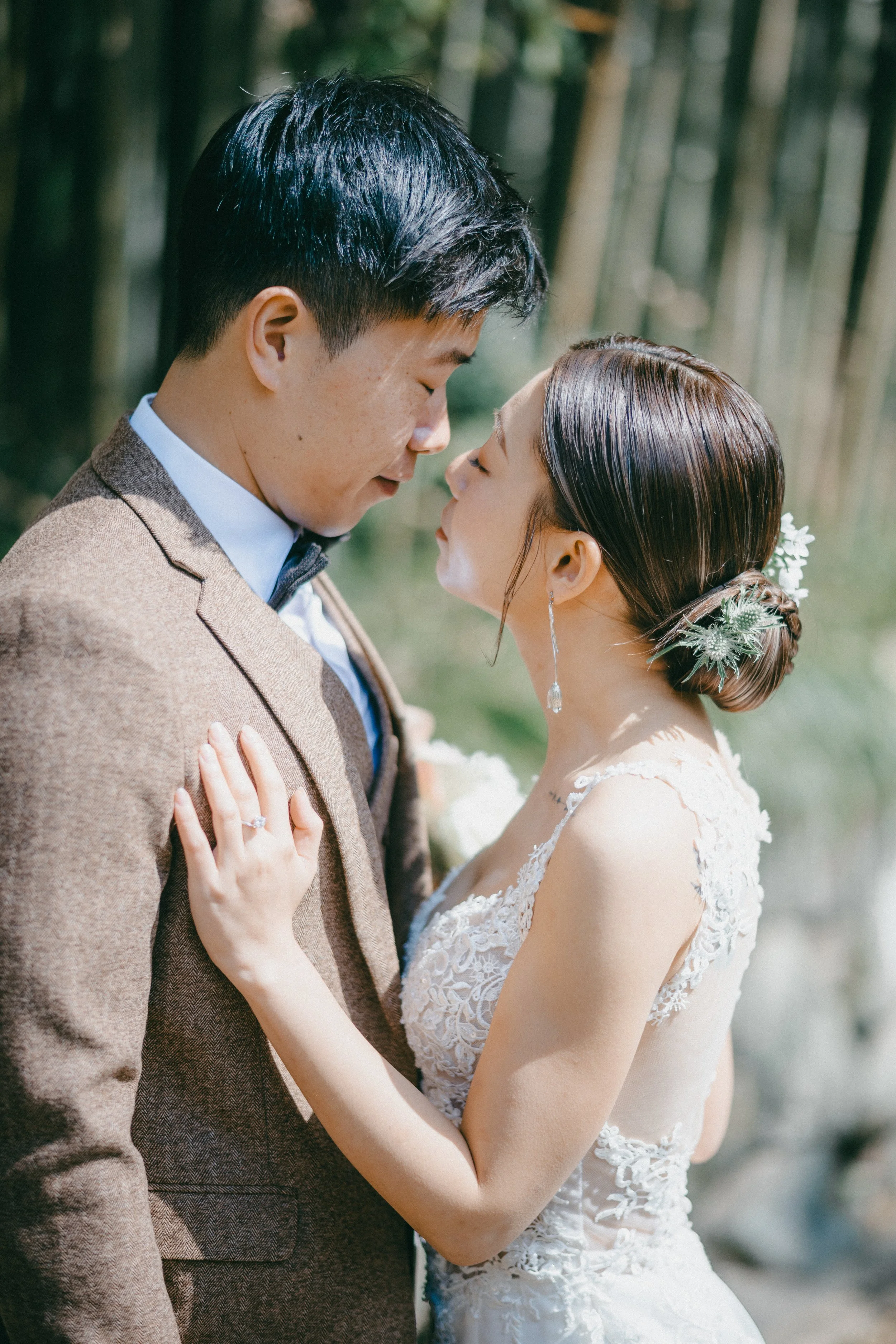 A bride and groom close together outdoors, with their foreheads touching and eyes closed, in a romantic pose. The bride wears a lace wedding gown and has her hair in a low bun with floral accessories. The groom wears a brown suit and has short dark h