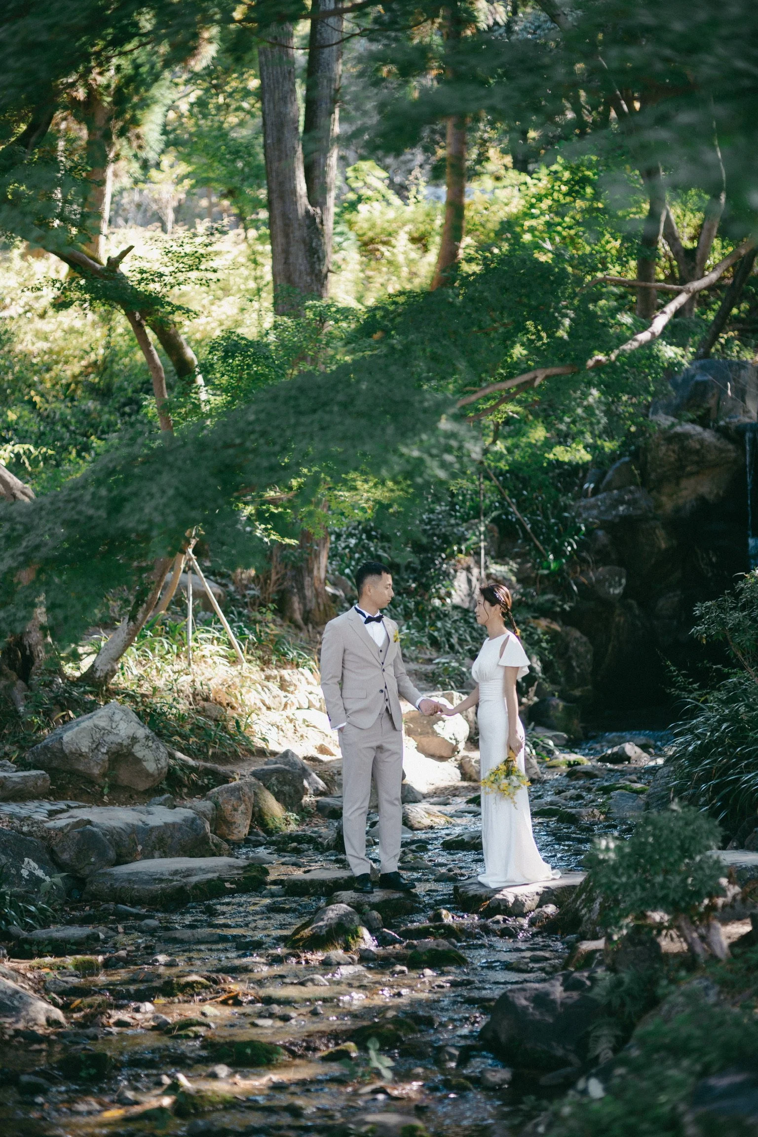 A bride and groom holding hands in a forested area with rocks and a small stream, dressed in wedding attire.