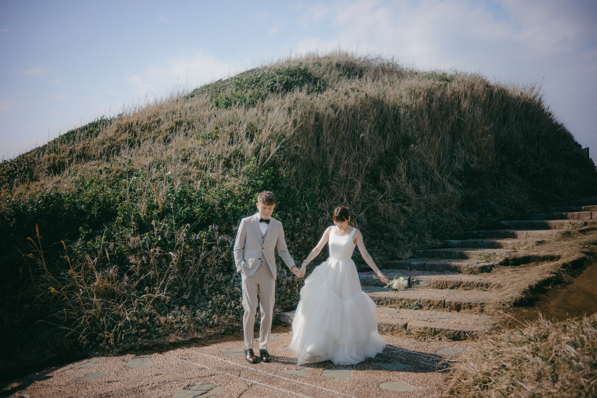 A bride and groom holding hands and walking down a stone path outdoors, with a grassy hill in the background.