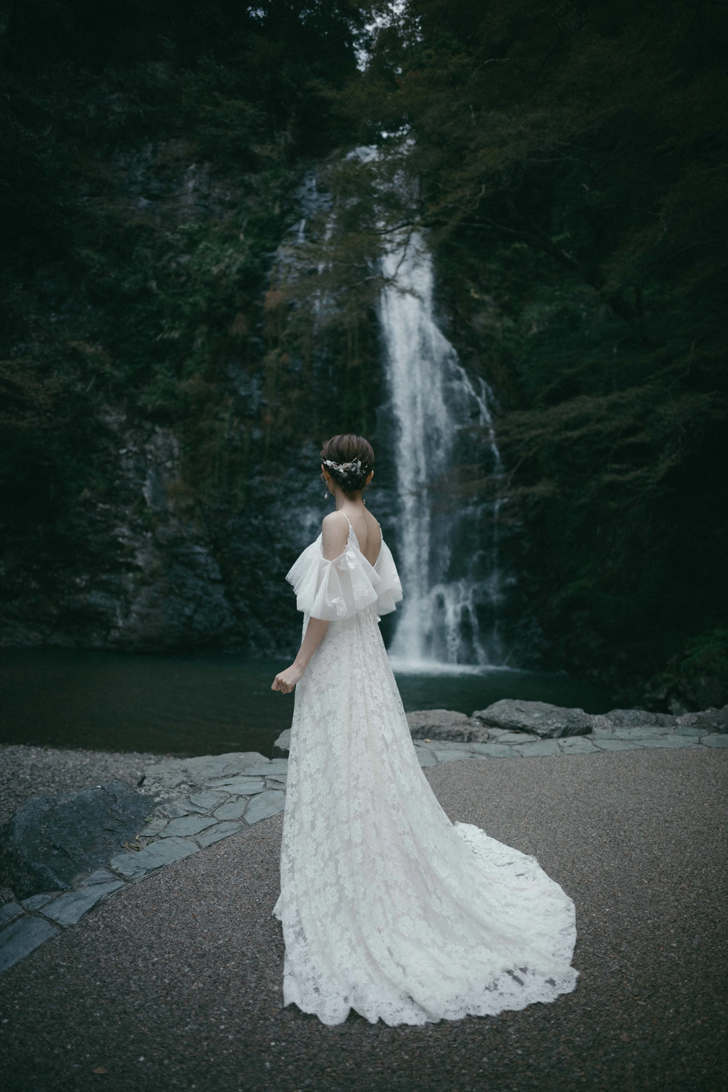 A woman in a white lace wedding dress stands outdoors near a waterfall surrounded by forest.