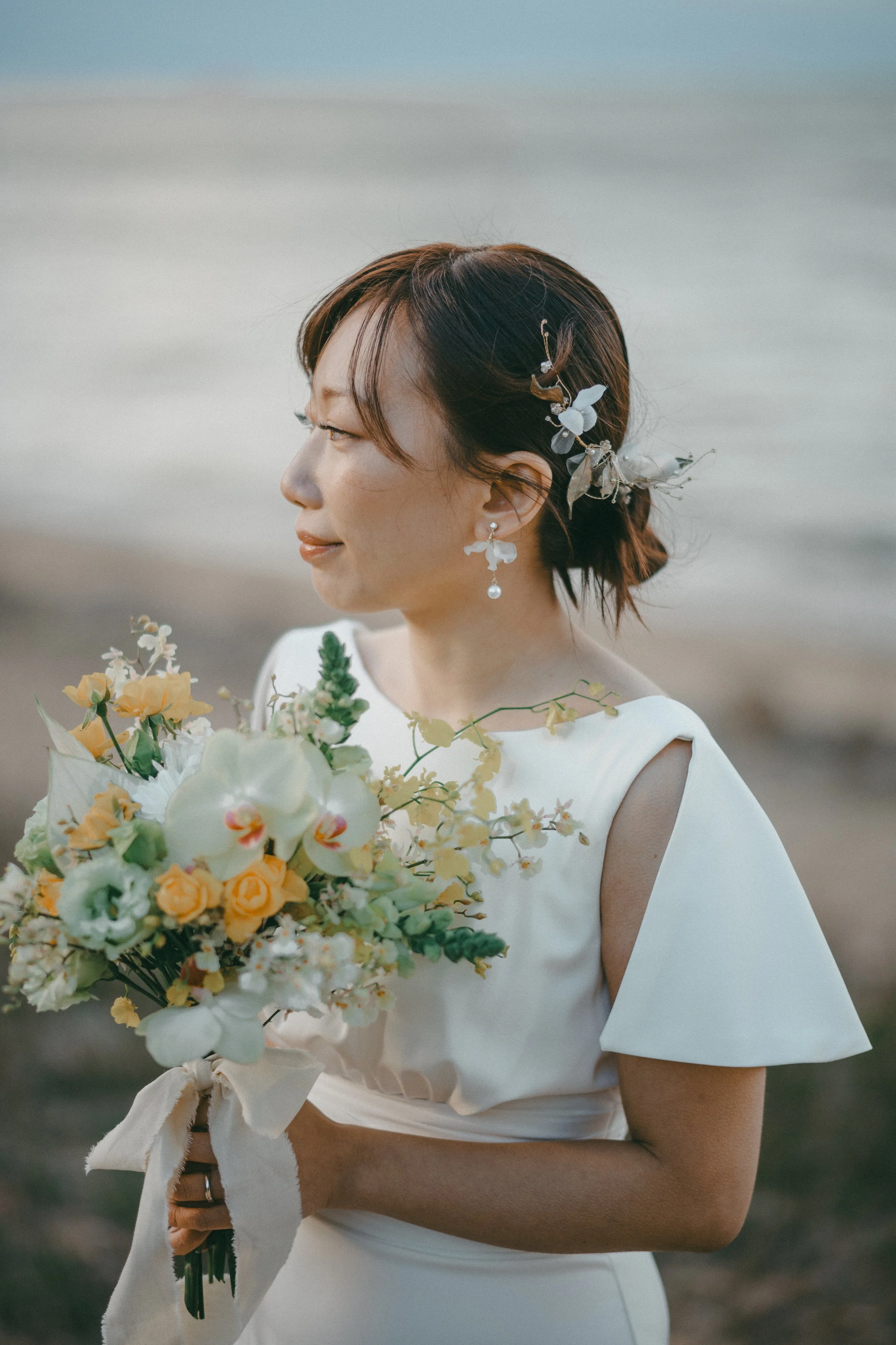 A bride in a white wedding dress stands on a beach, holding a bouquet of white and yellow flowers, with her hair adorned with floral accessories.