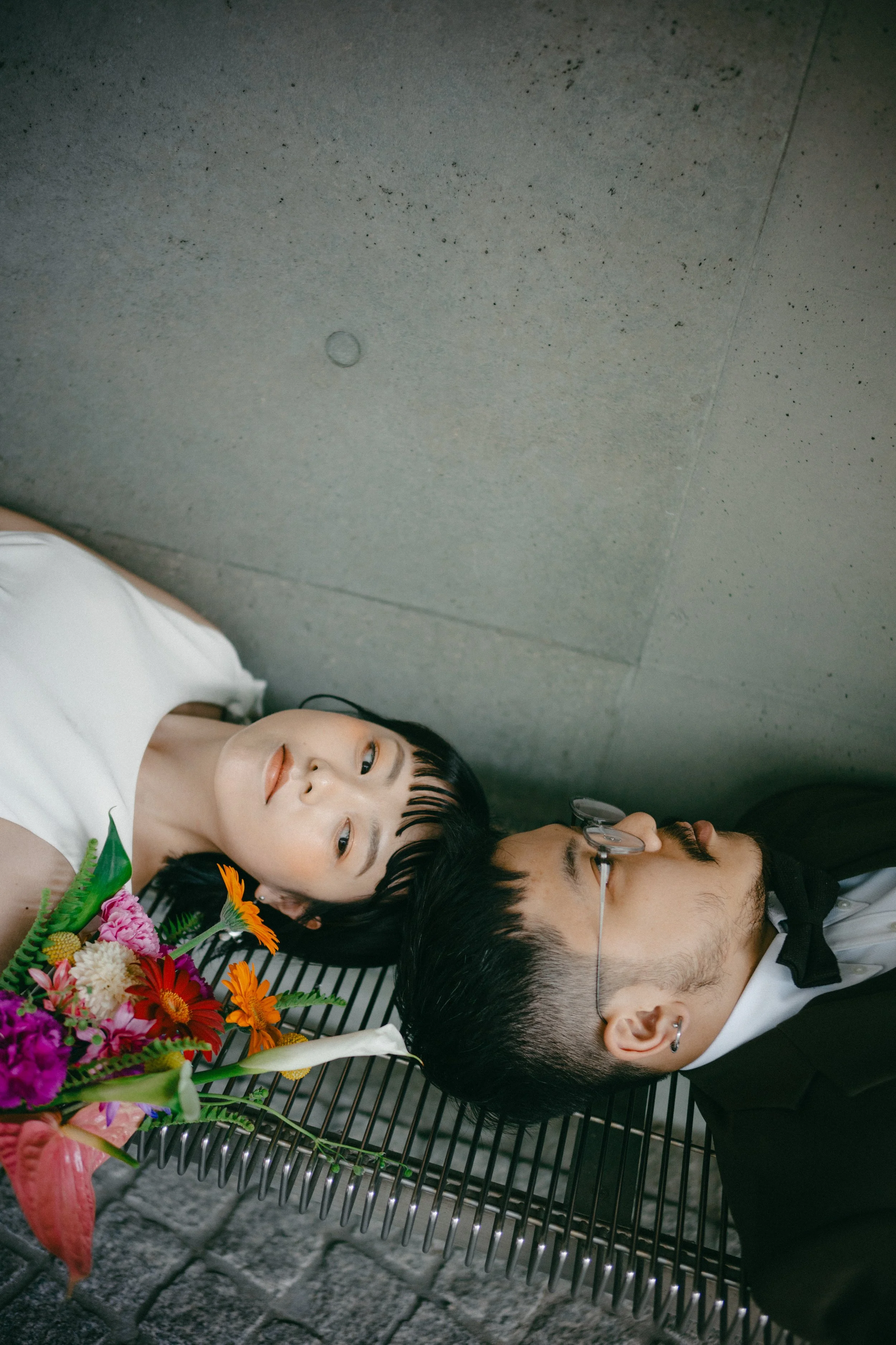 A young woman and a young man with glasses lying on a black metal bench with their heads together, surrounded by colorful flowers against a concrete wall.