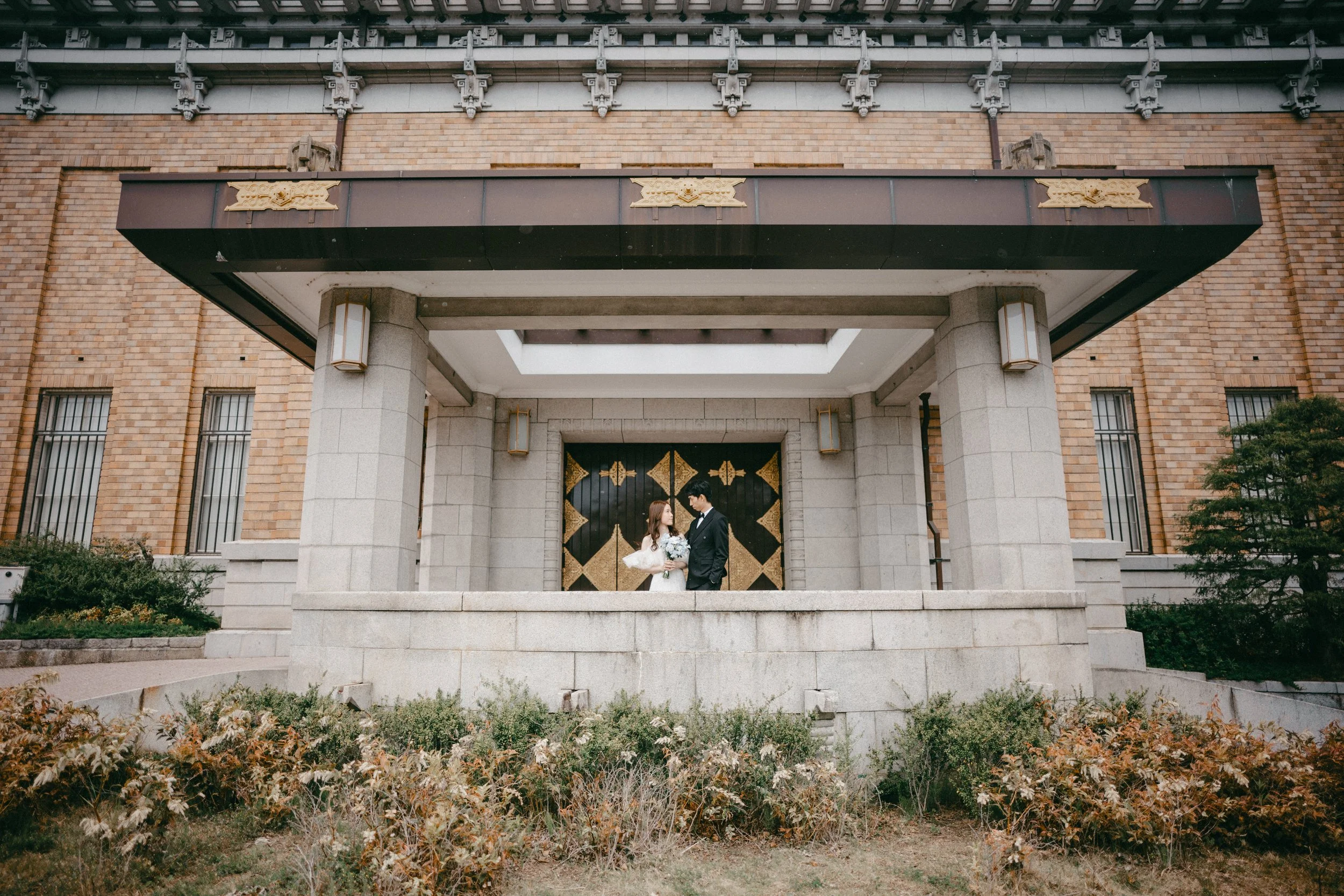 A bride and groom standing under a covered porch in front of a large black door with gold accents. The building has brick walls, lantern-style lights, and a small garden in the foreground.