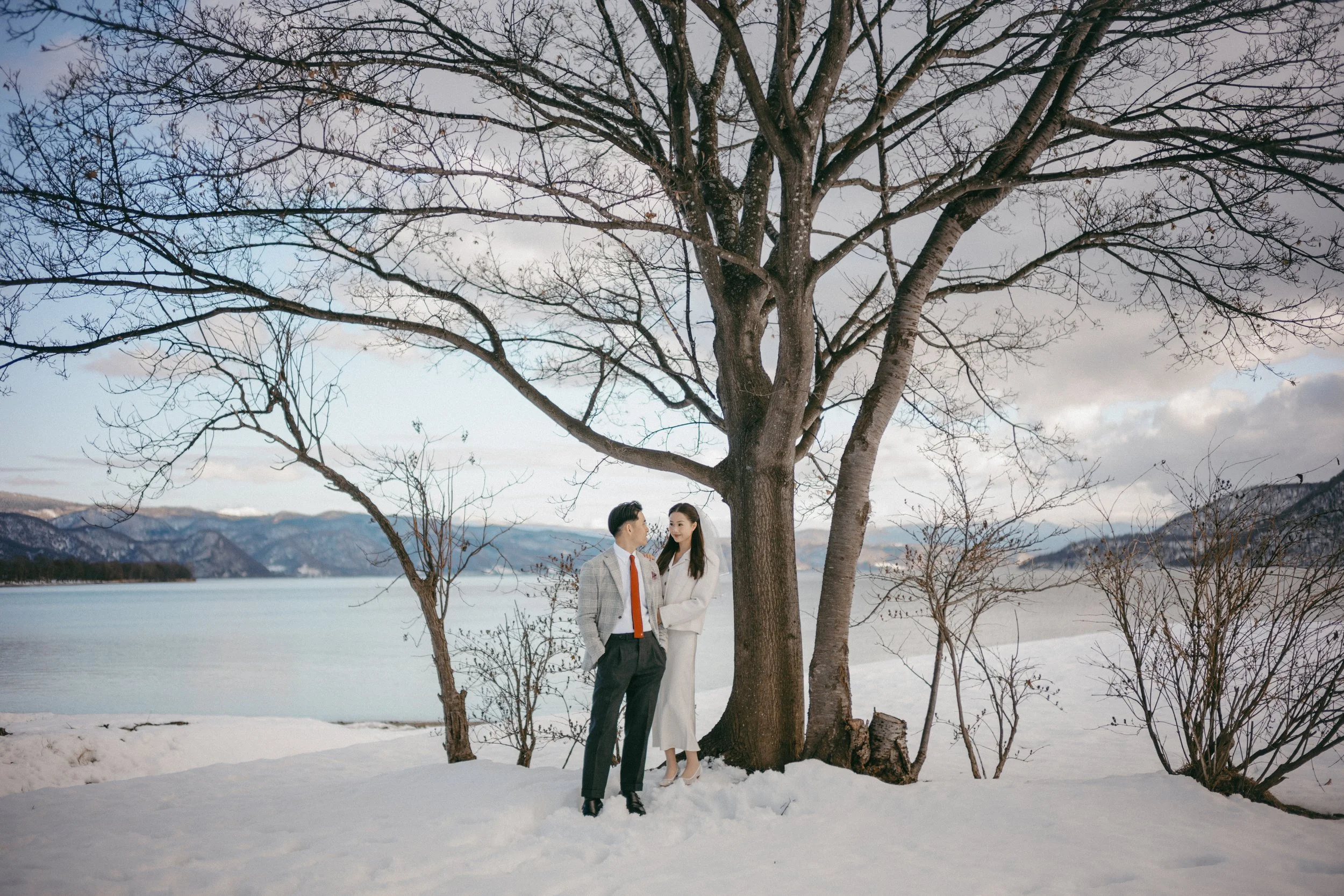 A man and woman dressed in formal winter attire standing near a large leafless tree on snow-covered ground by a lake, with mountains in the background under a cloudy sky.