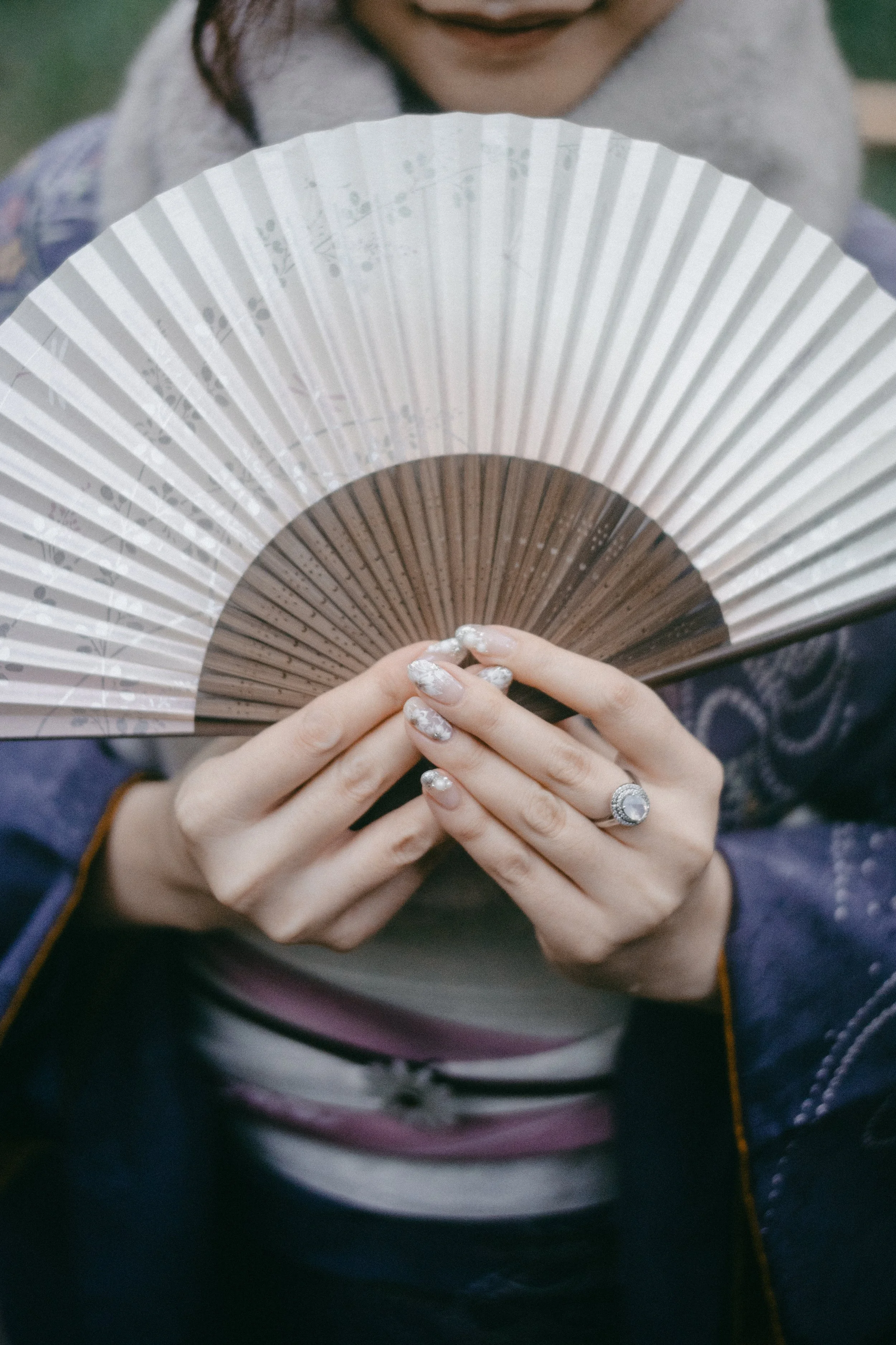 Person holding a folding fan with painted nails and a large ring on their finger.