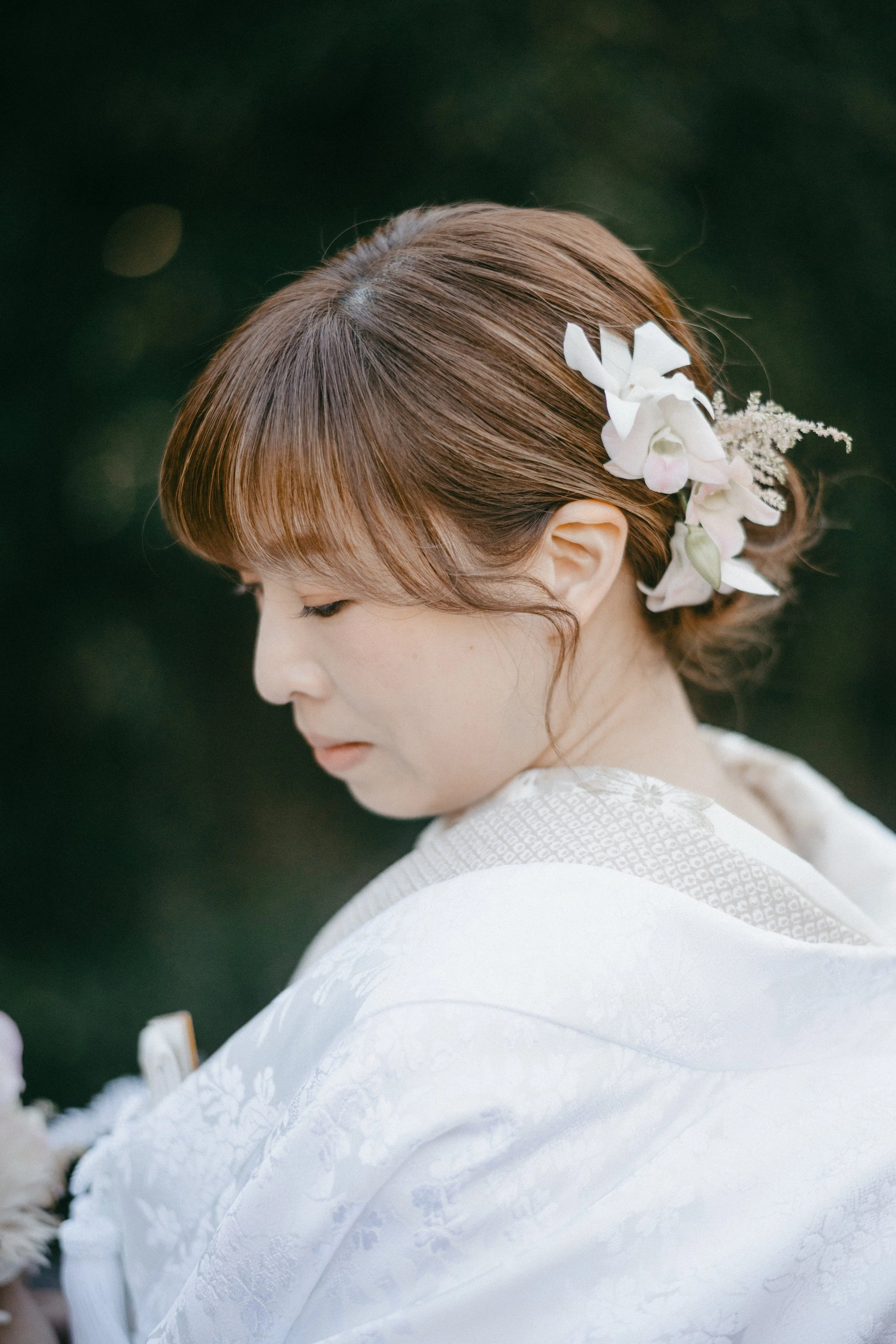 A young woman with brown hair styled with flowers, wearing a traditional white kimono, with a serene expression, against a dark, out-of-focus background.