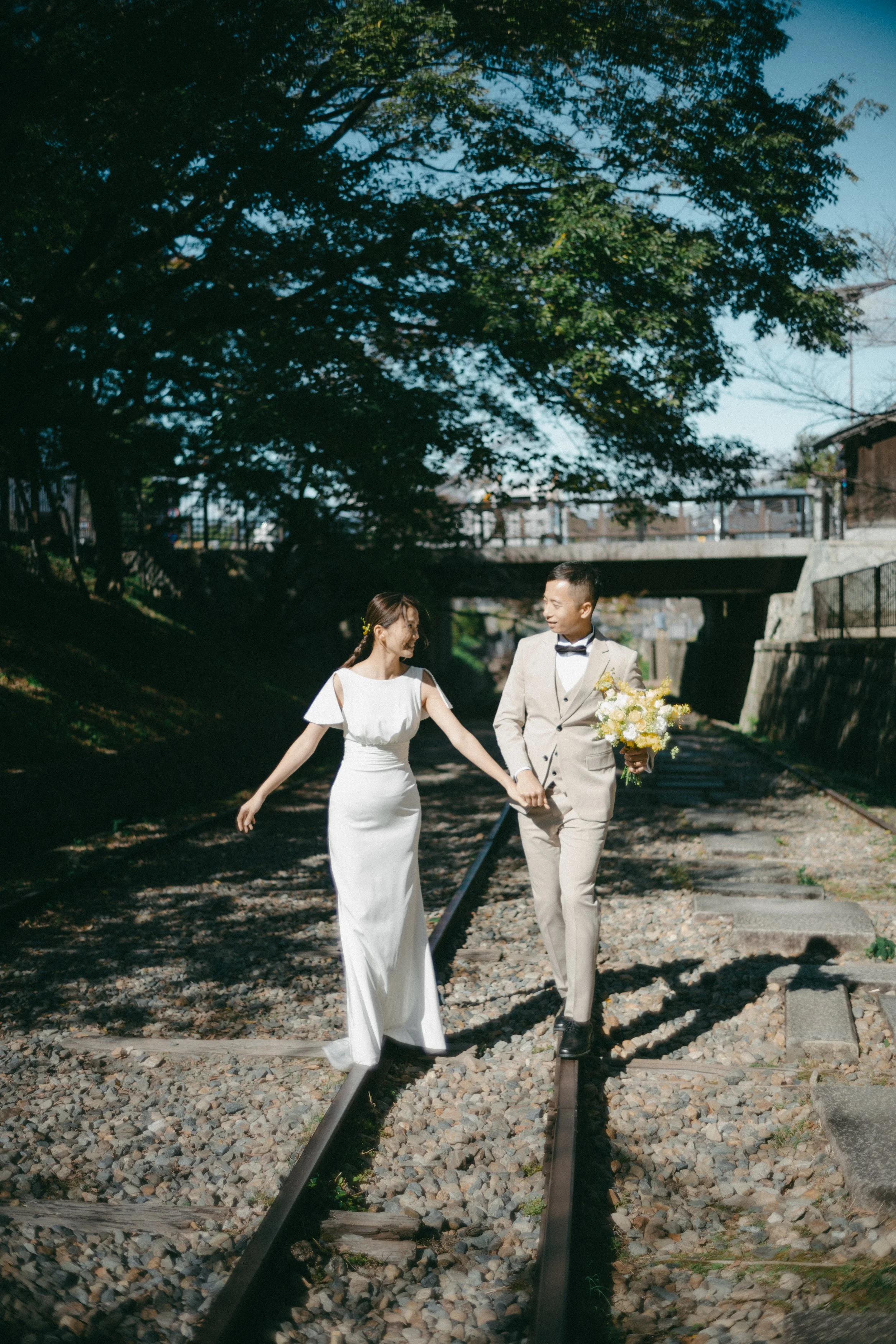 A bride and groom walking hand in hand on railroad tracks outdoors during daytime, with trees and a bridge in the background.
