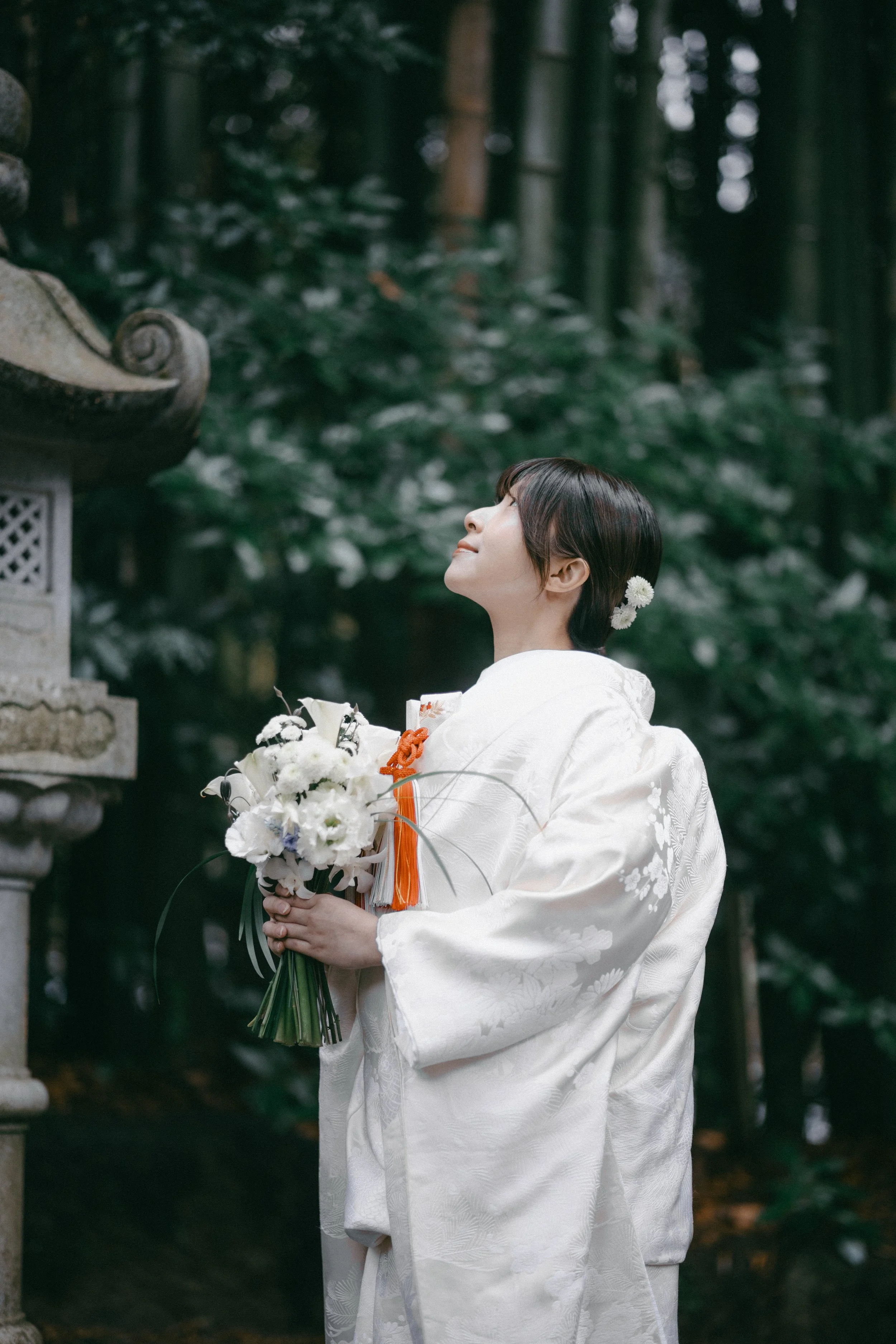 A woman in traditional Japanese wedding kimono holding a bouquet of white flowers, standing in a forest near a stone lantern, looking upwards with a serene expression.