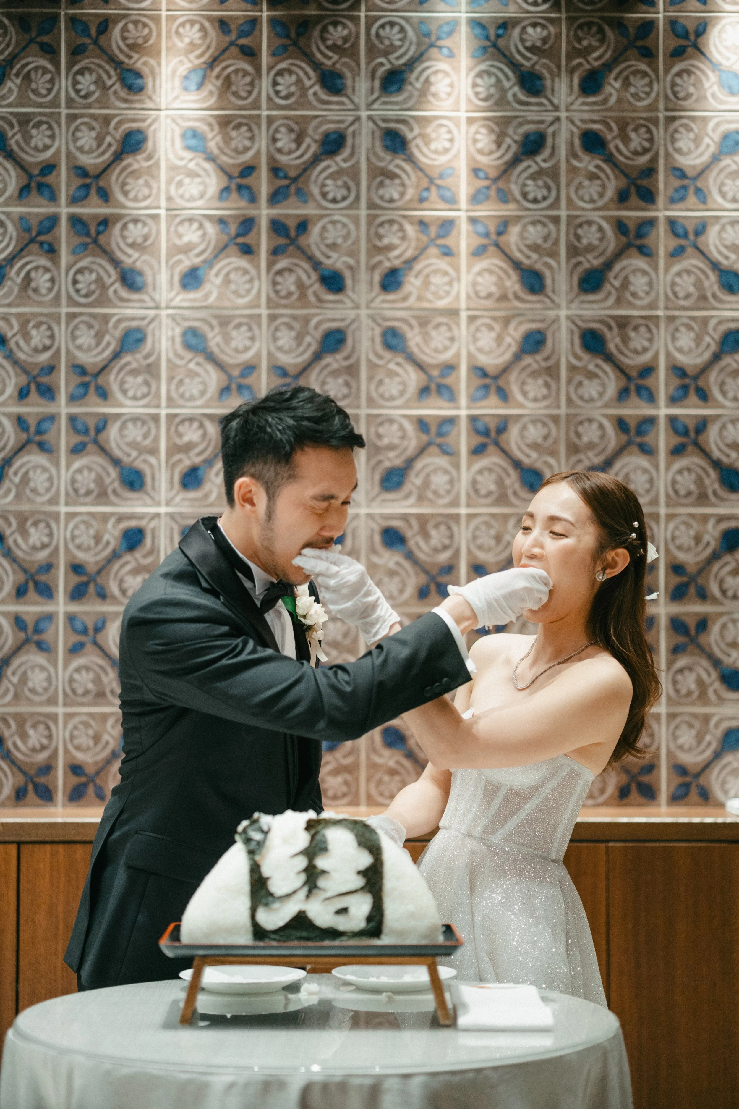 A bride and groom celebrate at their wedding with a traditional cake-cutting ceremony, with a decorative tiled wall in the background.