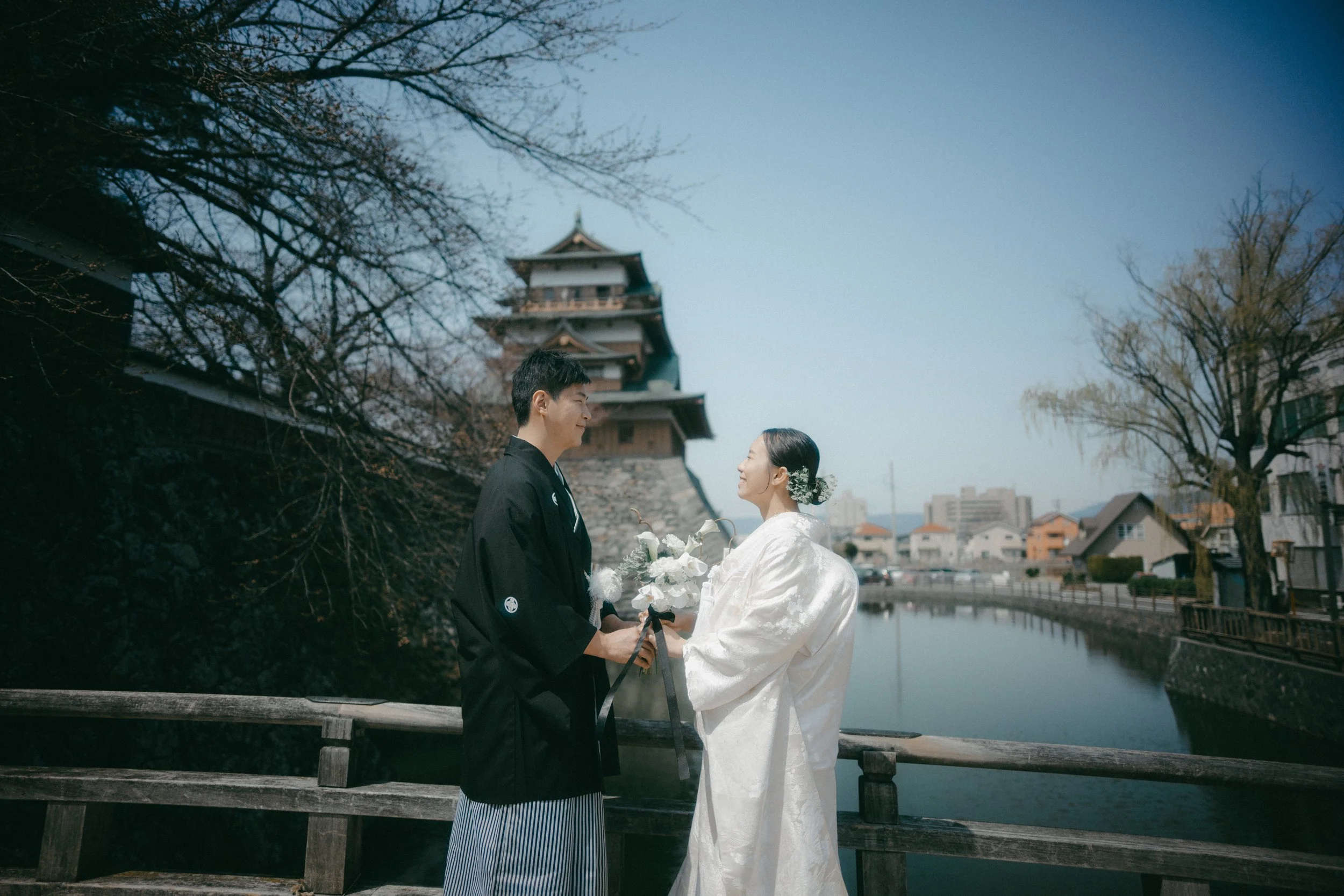 A bride and groom in traditional Japanese wedding attire stand on a bridge, holding flowers and facing each other, with a historic Japanese castle and river in the background.