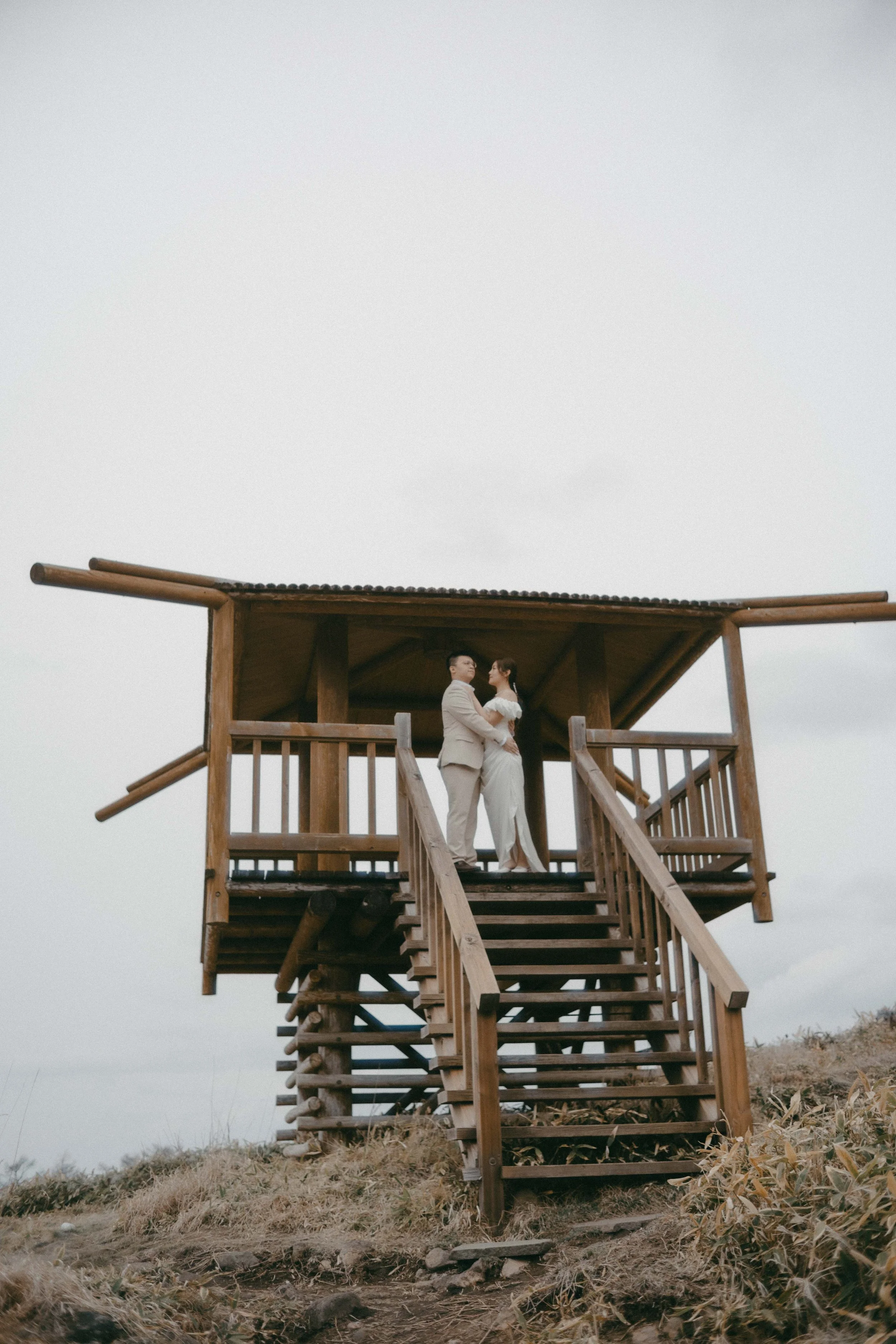 A couple dressed in wedding attire standing on a wooden observation deck, holding hands and looking at each other, with an overcast sky in the background.