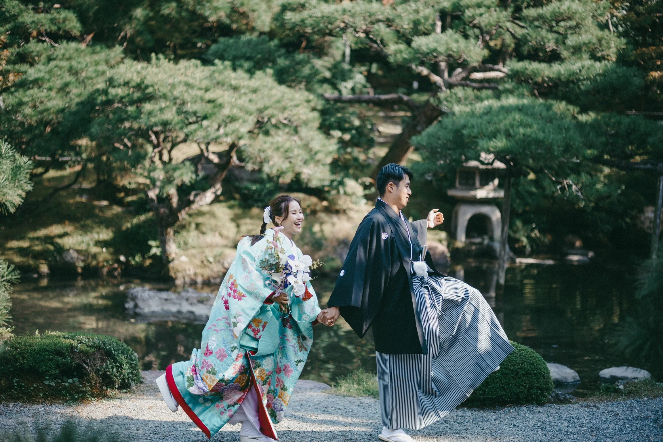A couple dressed in traditional Japanese wedding attire walking by a pond in a Japanese garden.