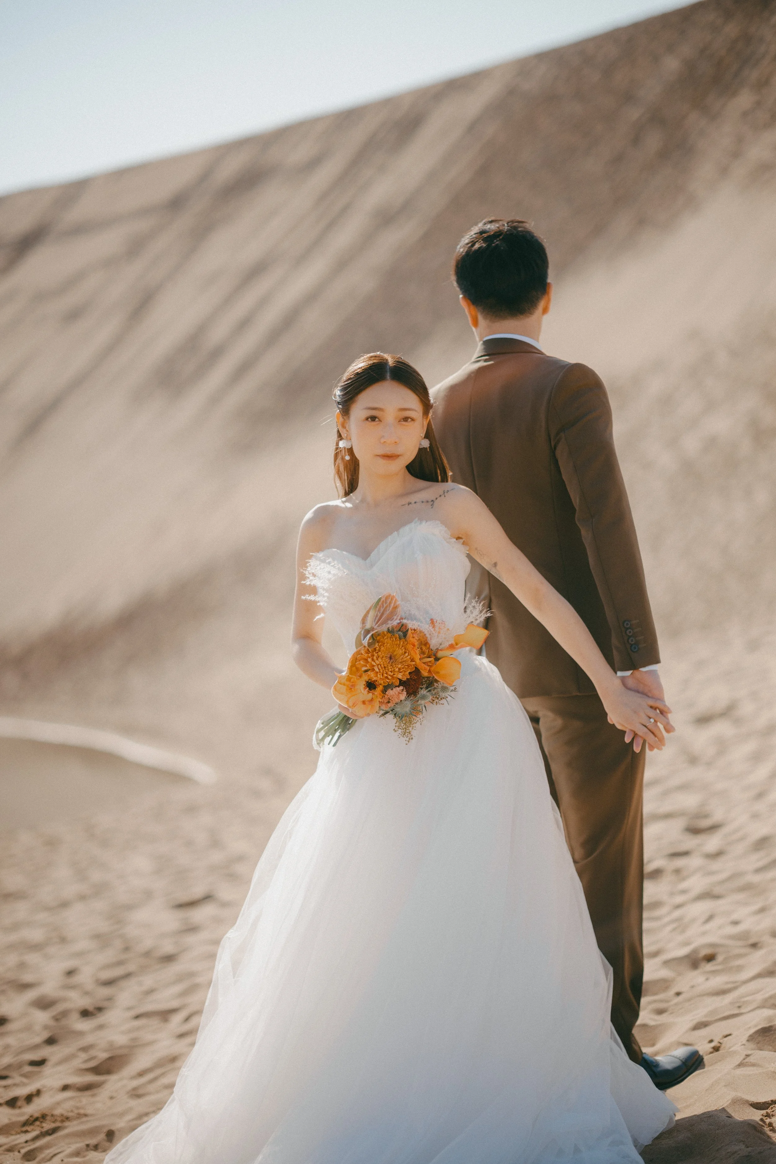 A bride in a white wedding gown holding a bouquet of orange flowers, standing in a desert with a groom dressed in a brown suit facing away in the background.