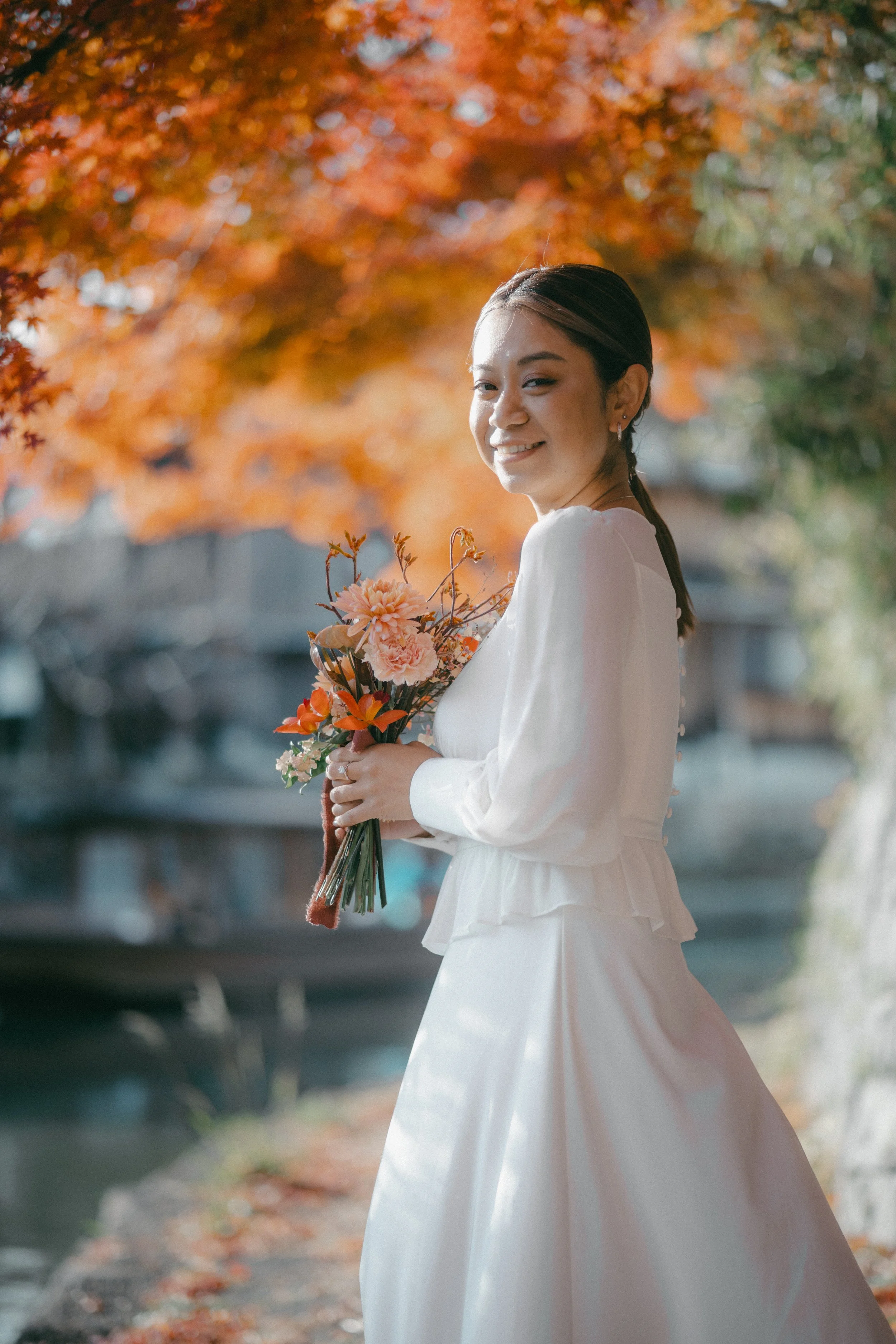 A woman in a white dress holding a bouquet of pink and orange flowers, standing outdoors with orange autumn leaves in the background.