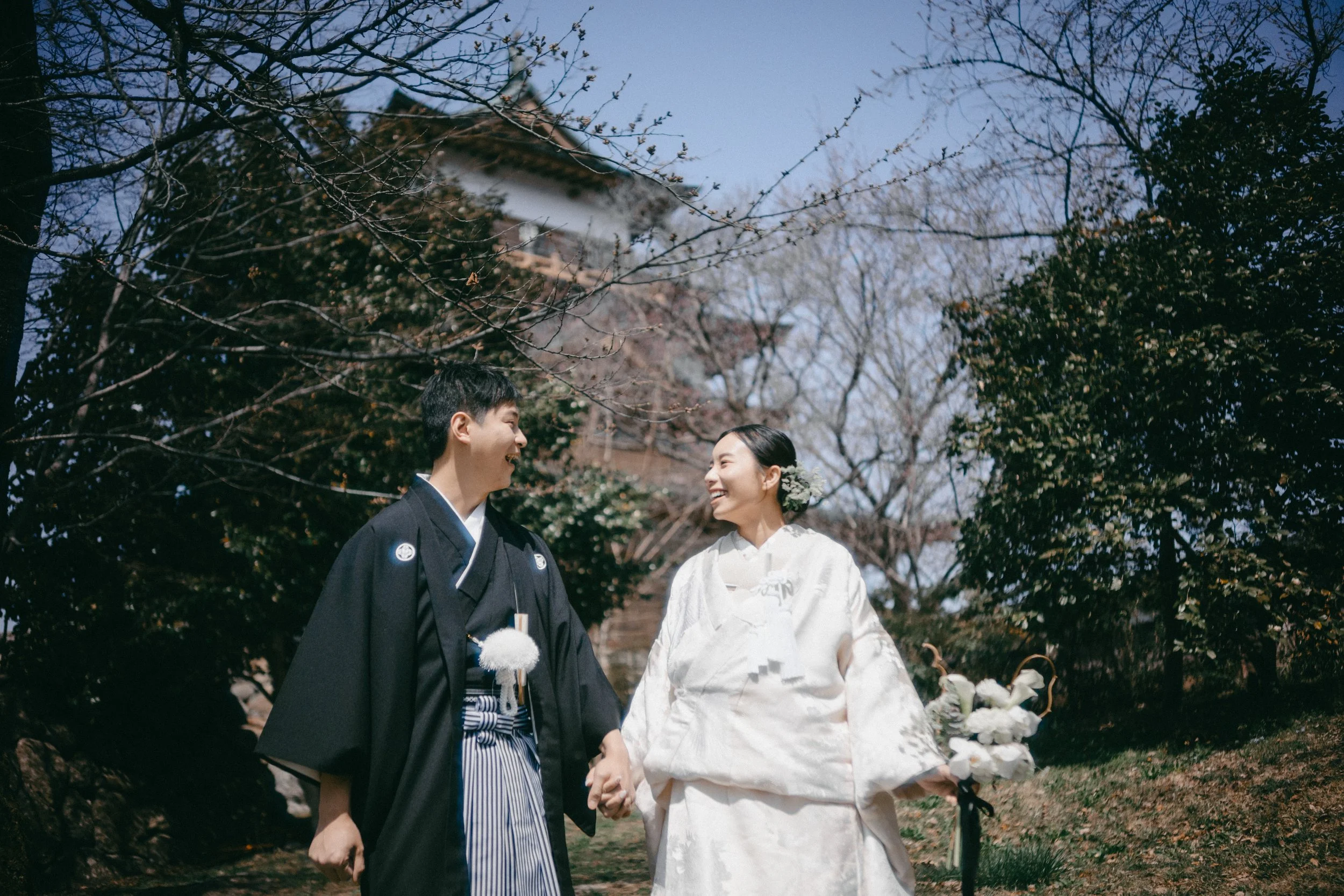 A newly married couple dressed in traditional Japanese wedding attire holding hands and smiling at each other outdoors. The groom wears a formal black kimono with a striped hakama, and the bride wears a white wedding kimono with a floral hair accesso