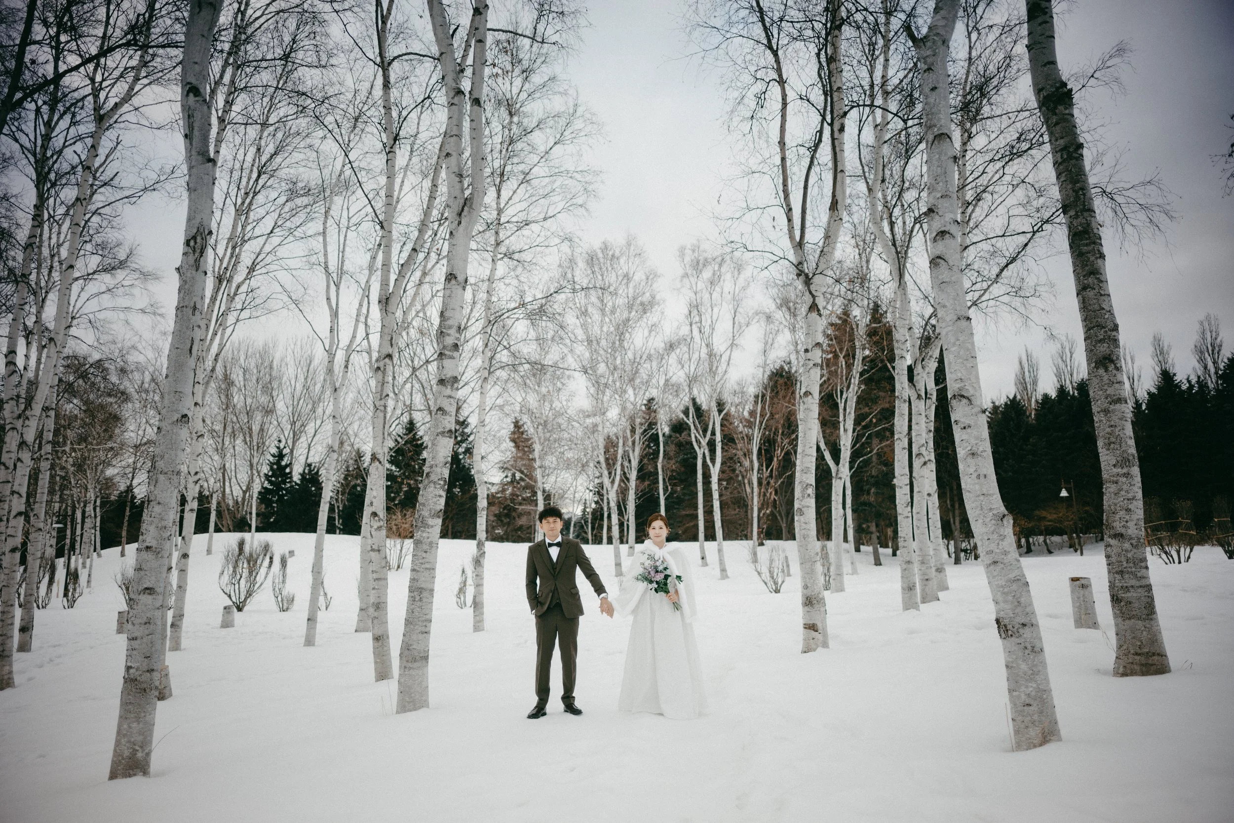 A bride and groom holding hands in a snowy forest with leafless trees