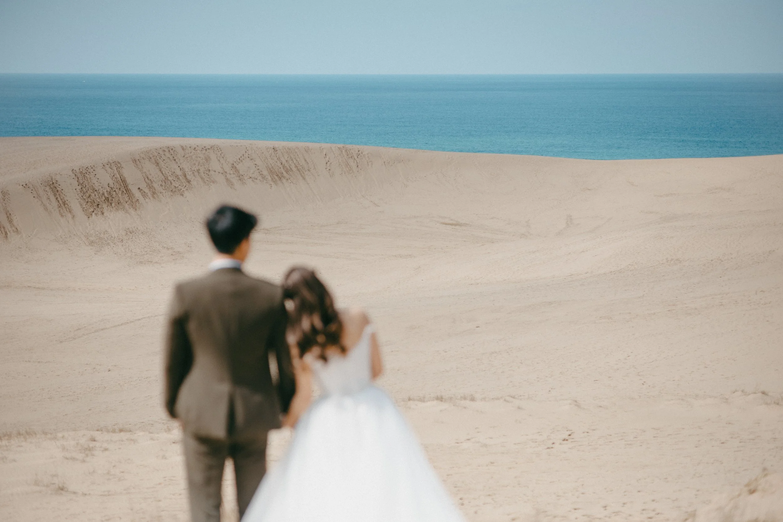 A couple in wedding attire walking on a sandy beach with the ocean in the background.