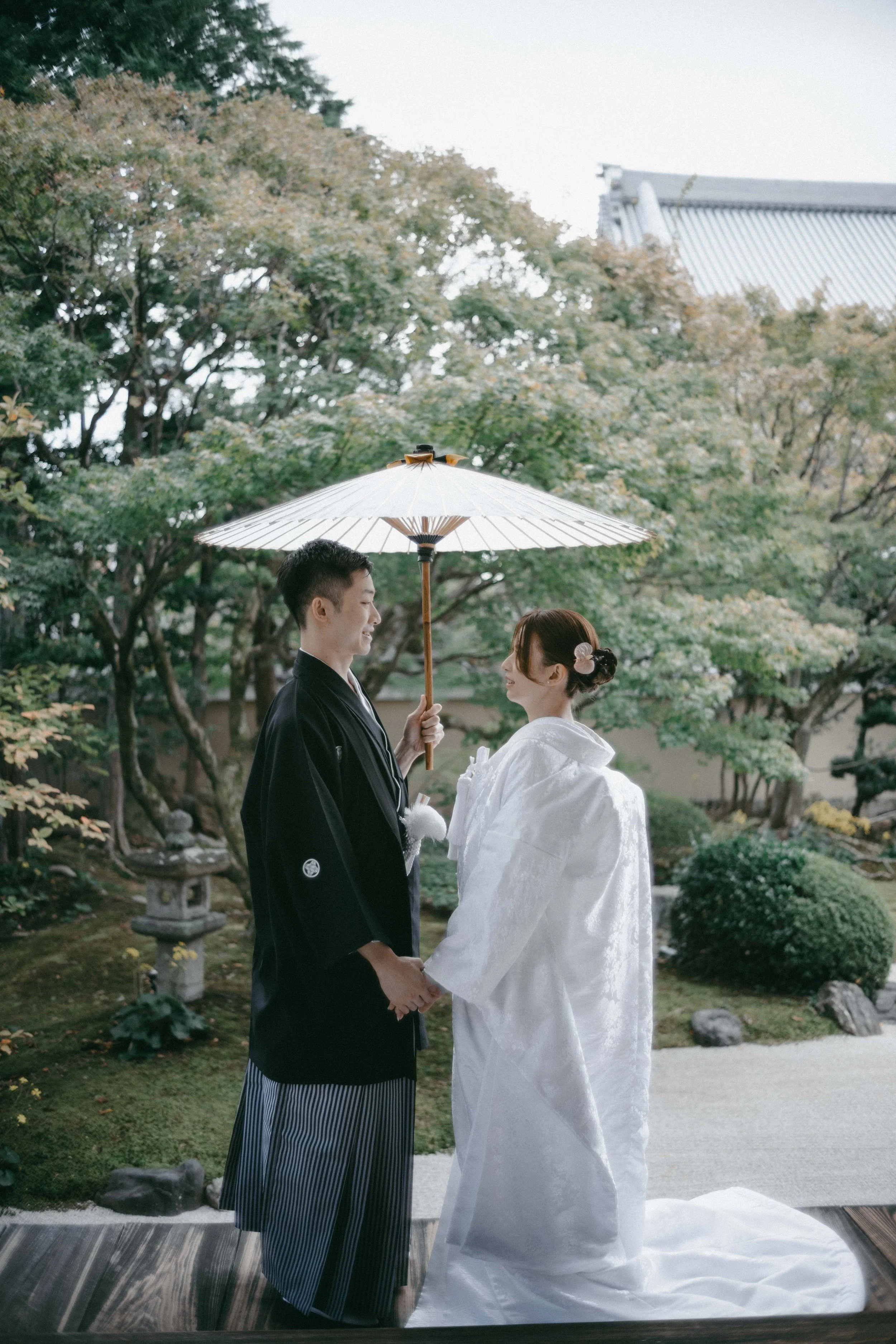 A couple dressed in traditional Japanese wedding attire holding hands and smiling at each other outdoors, with the man holding a paper umbrella over them. The background features lush greenery and a traditional Japanese building.