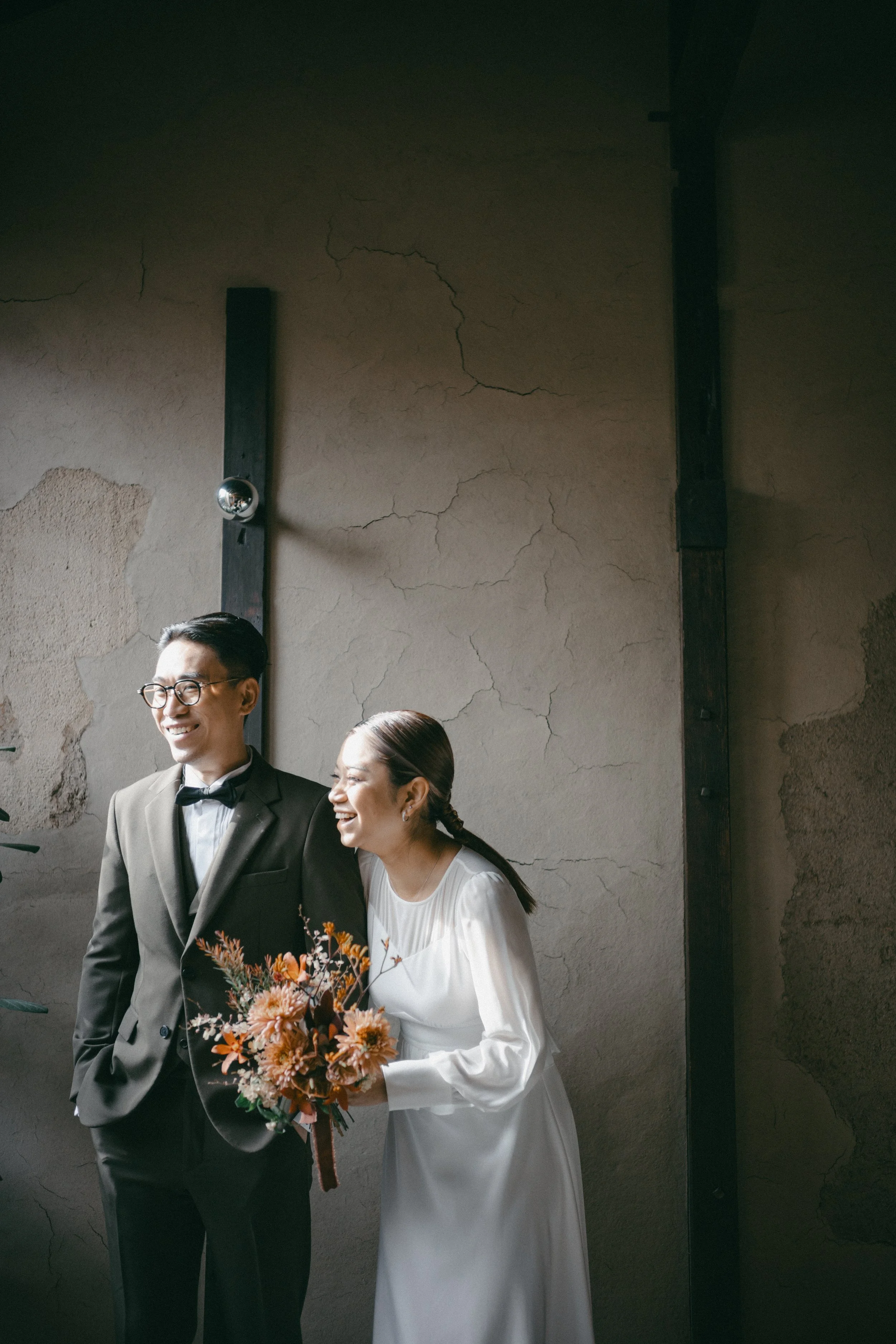 A happy couple, the man in a tuxedo and the woman in a white dress holding a bouquet, standing indoors near a textured wall with cracks, smiling and laughing together.