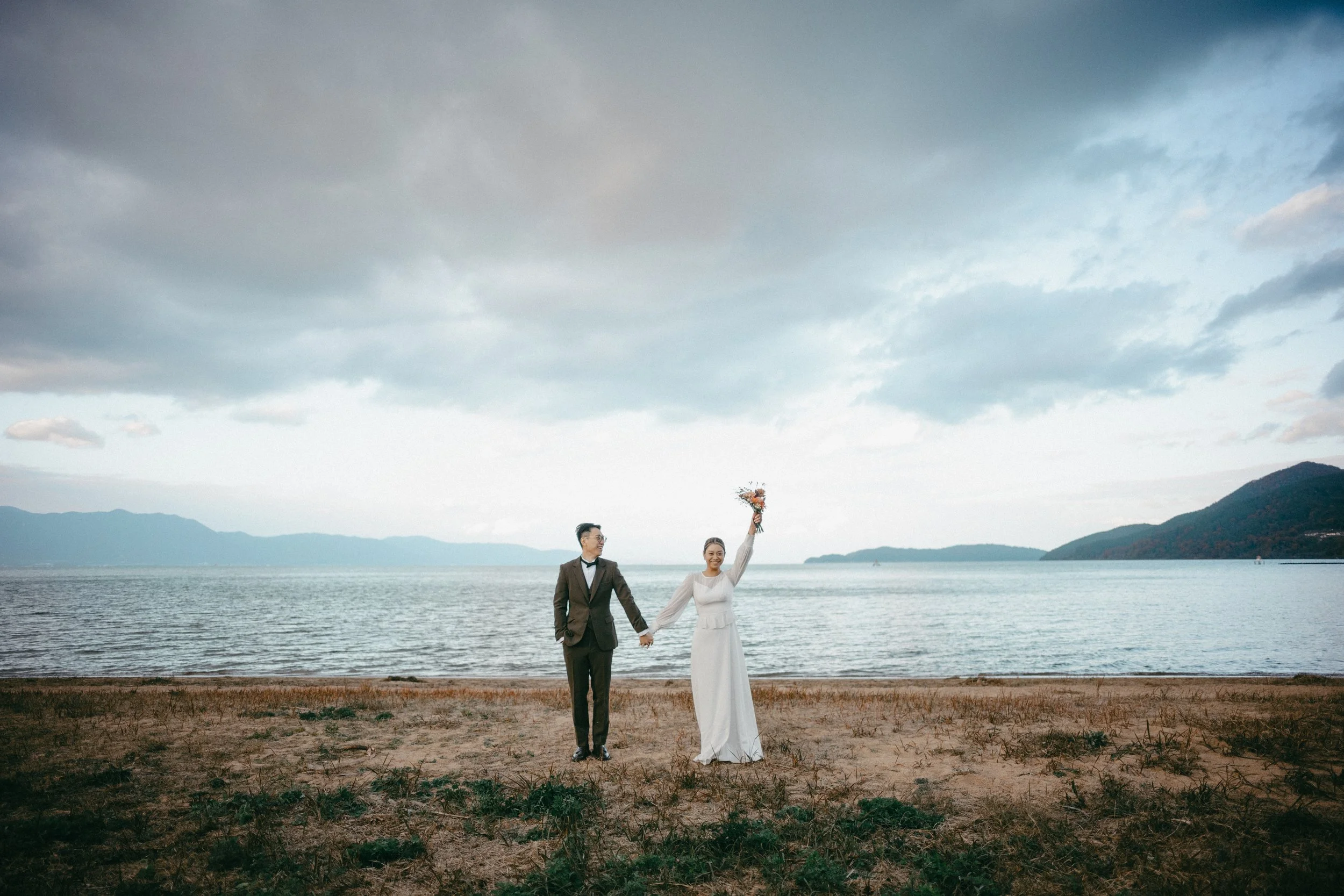 A newlywed couple holding hands on a beach, with the bride raising a bouquet, overlooking a body of water and mountains under a cloudy sky.